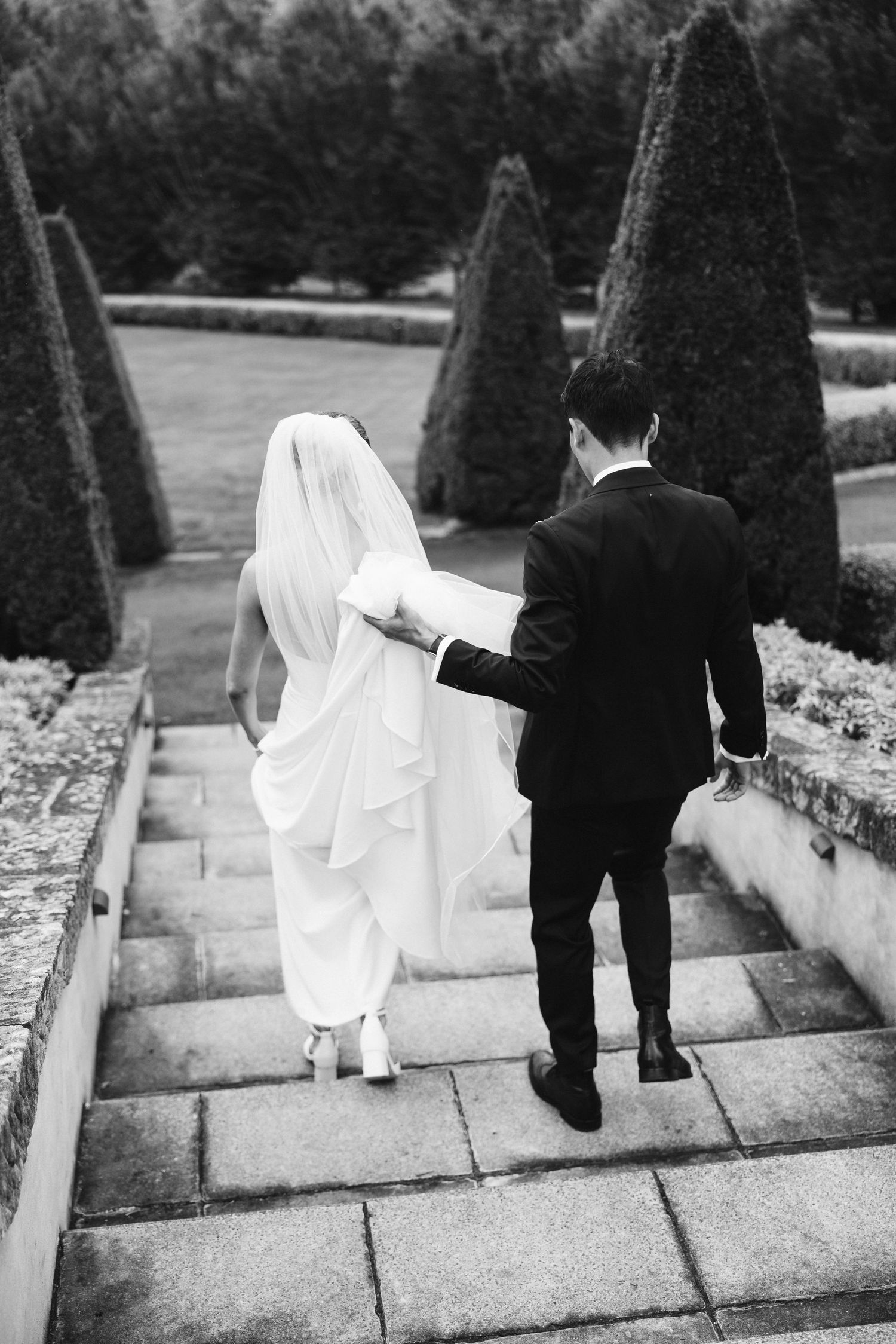 Black and white photo of newlyweds descending stone steps together in formal garden setting.