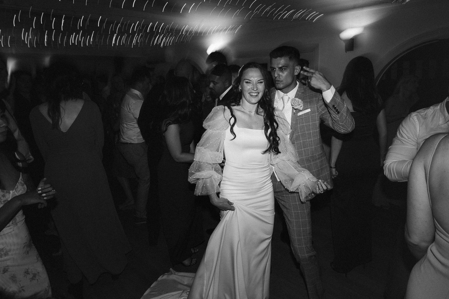 Black and white photo of a bride and groom dancing joyfully at their wedding reception surrounded by guests.