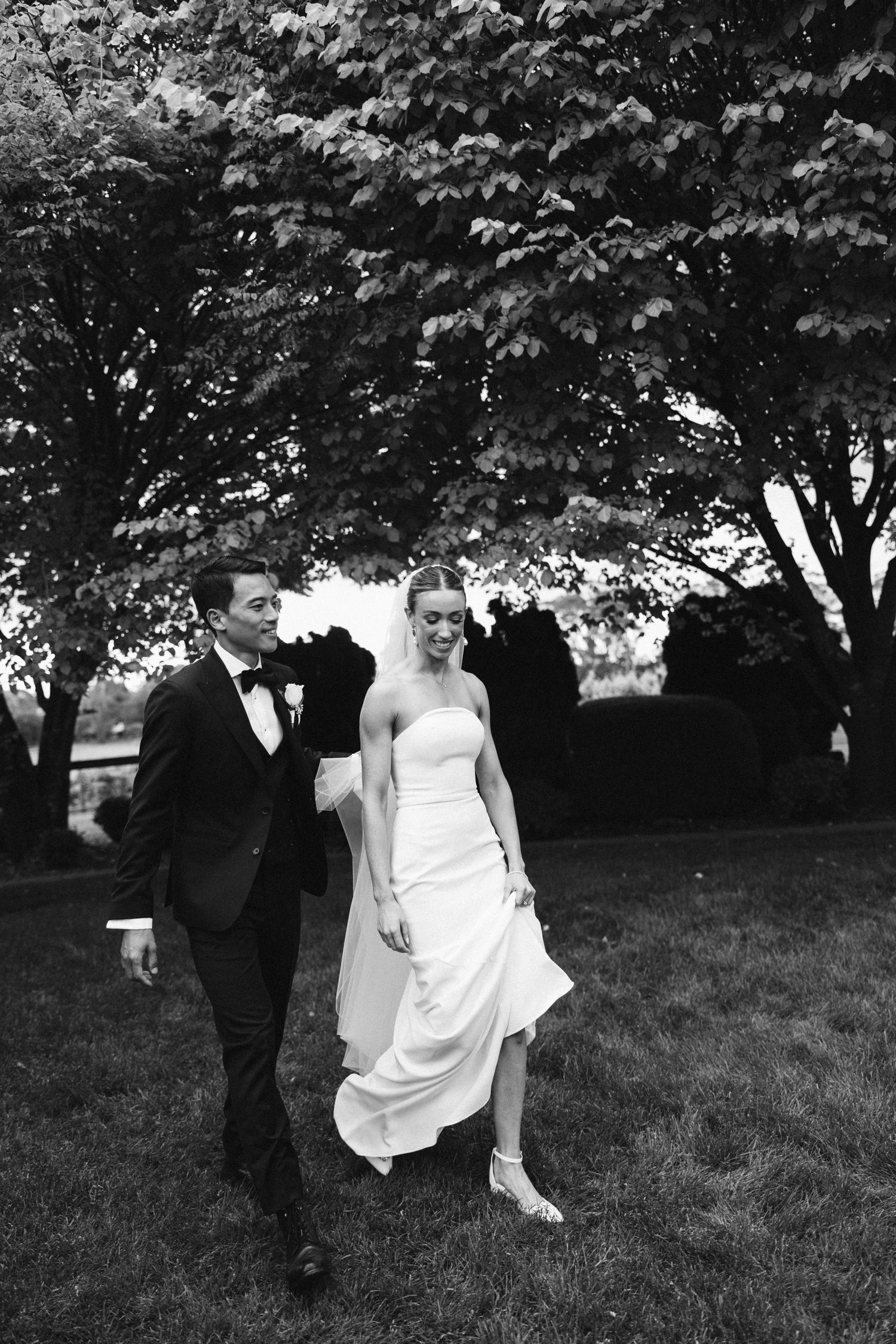 Wedding couple strolling on grass under leafy trees in black and white photography.
