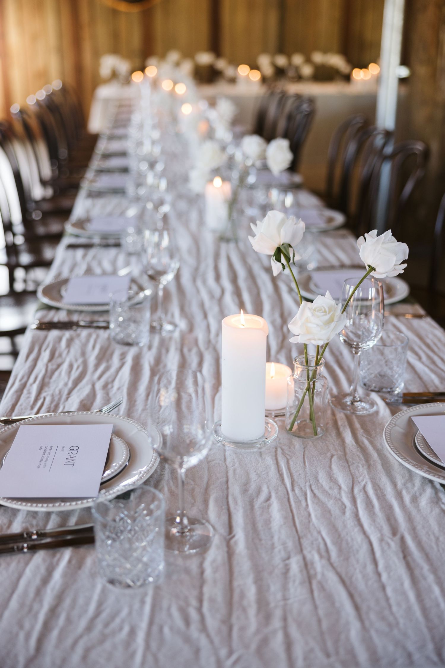 Elegant table setting with white linens, candles, and simple flower arrangements in glass vases.