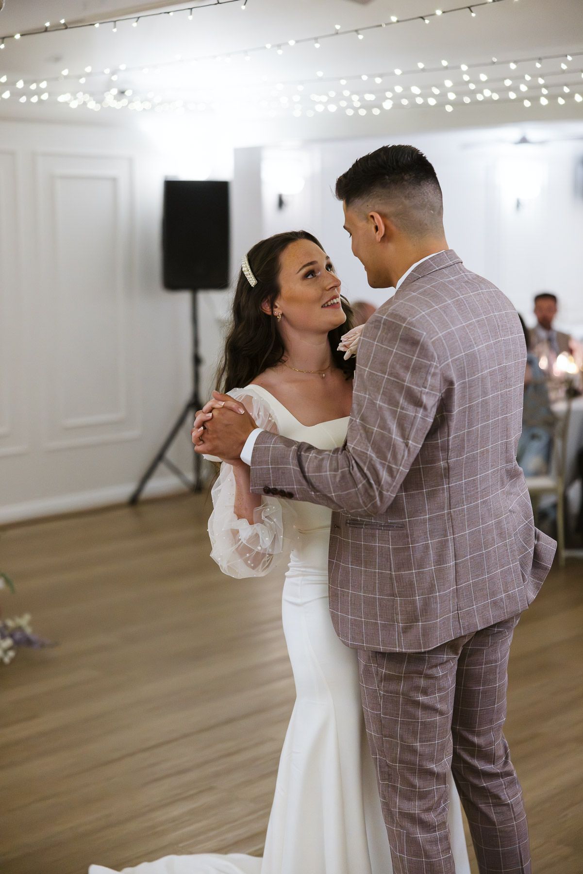 Bride and groom share their first dance, she in a white gown, he in a plaid suit, under fairy lights.