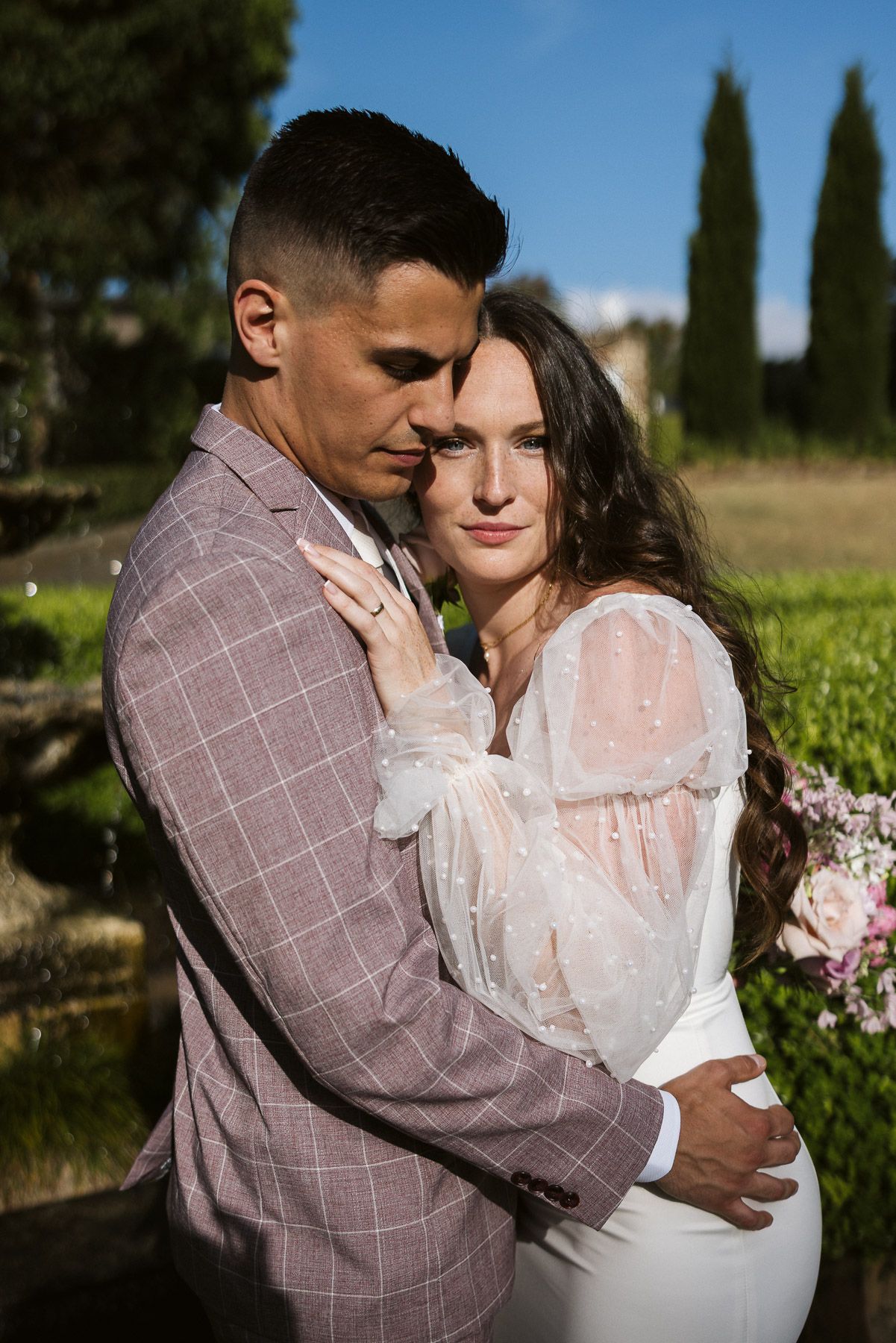 Romantic couple in wedding attire embracing outdoors, groom in plaid suit, bride in white sheer dress, garden backdrop.