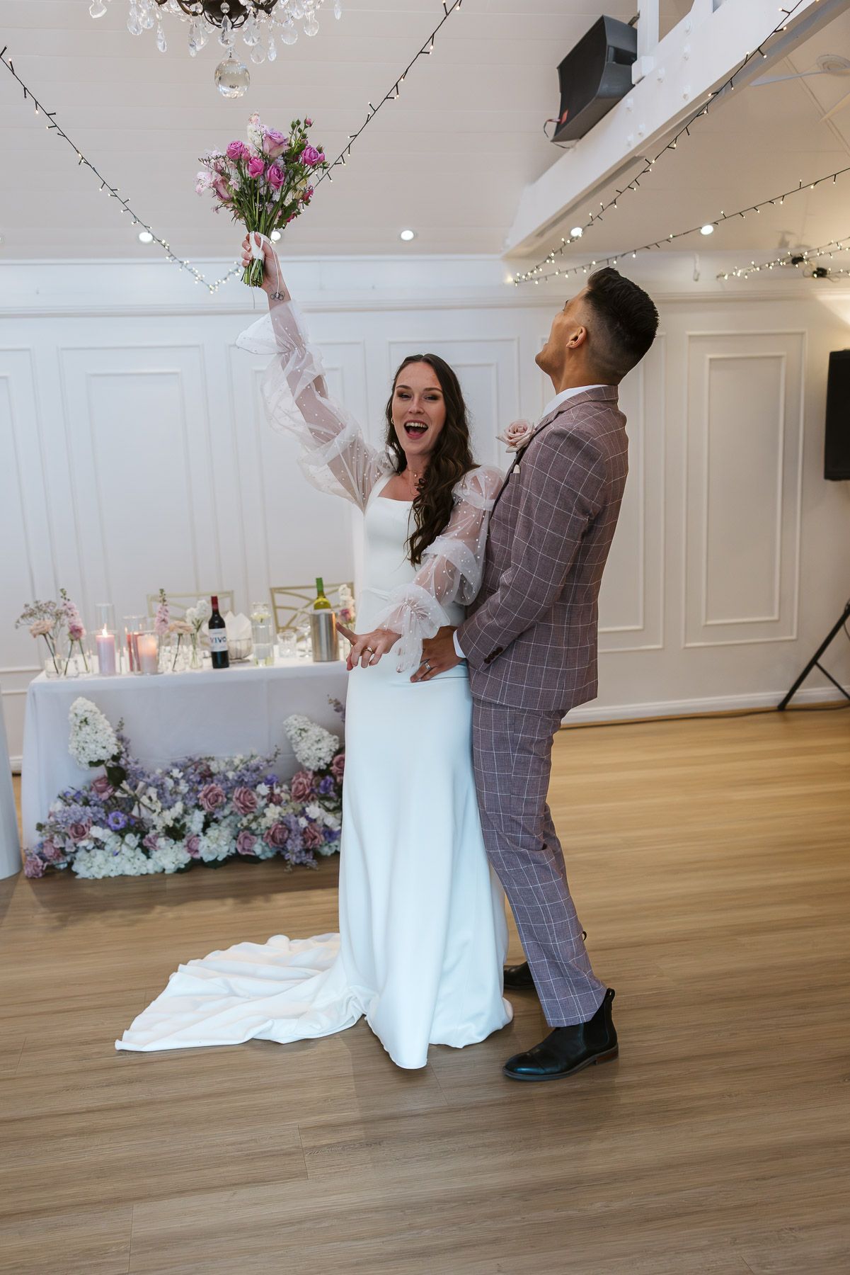Joyful bride in white gown raises bouquet while groom in plaid suit smiles during wedding reception dance.
