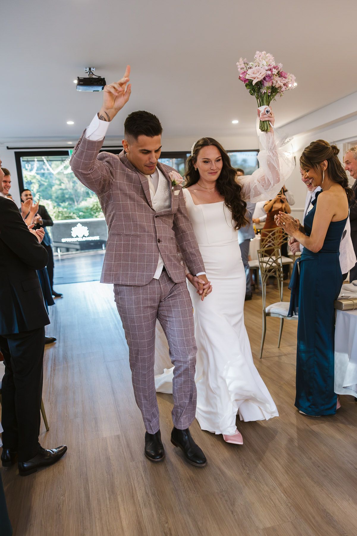 Joyful newlyweds walk down the aisle, groom in plaid suit raising hand, bride in white gown holding pink bouquet.