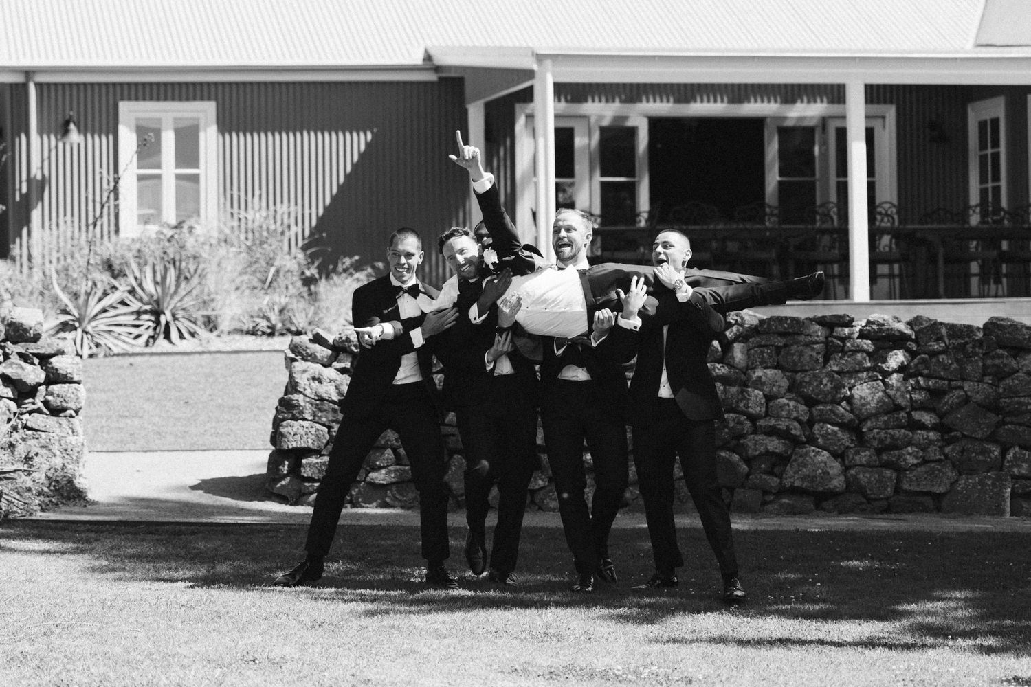 A black and white photo of groomsmen posing together on the porch of a rustic house with stone wall.