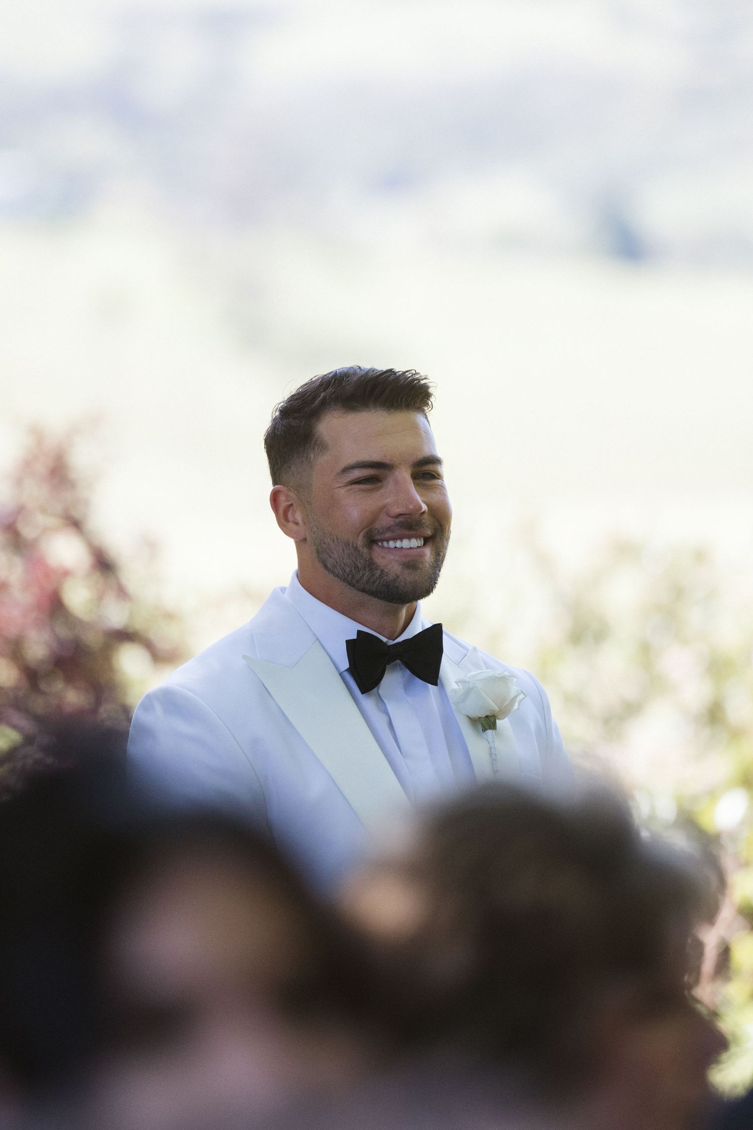 Groom in white tuxedo jacket and bow tie smiles during outdoor ceremony.
