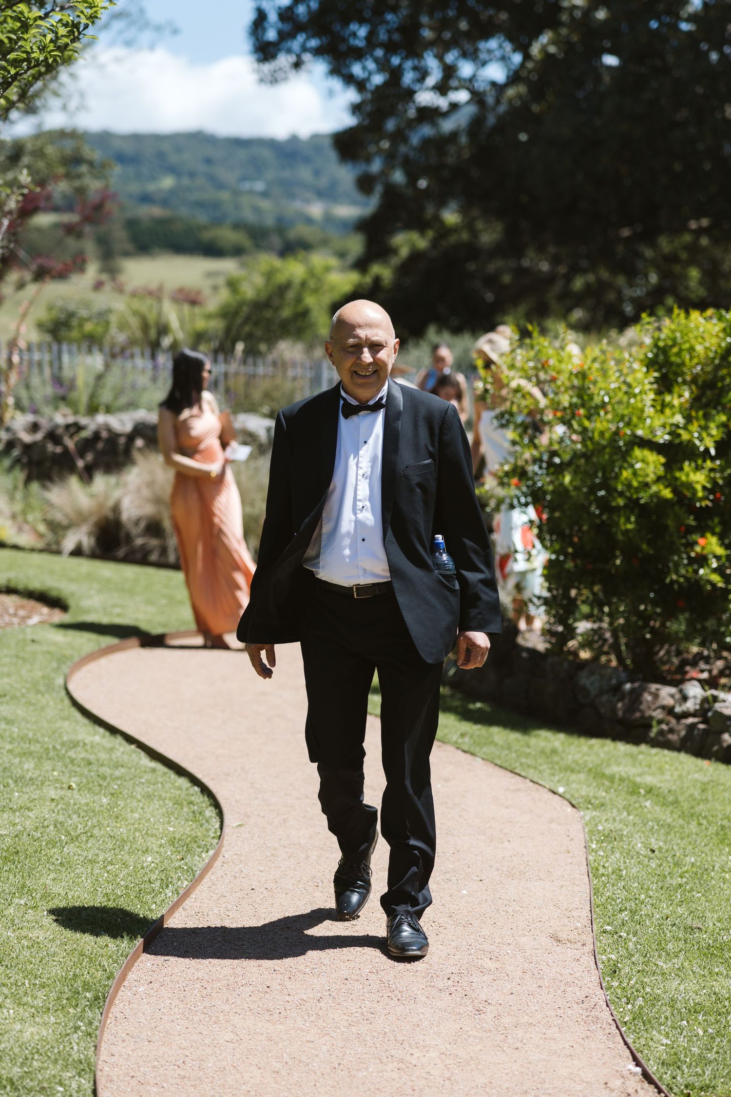 Formally dressed guest walks along garden path at outdoor event on sunny day.