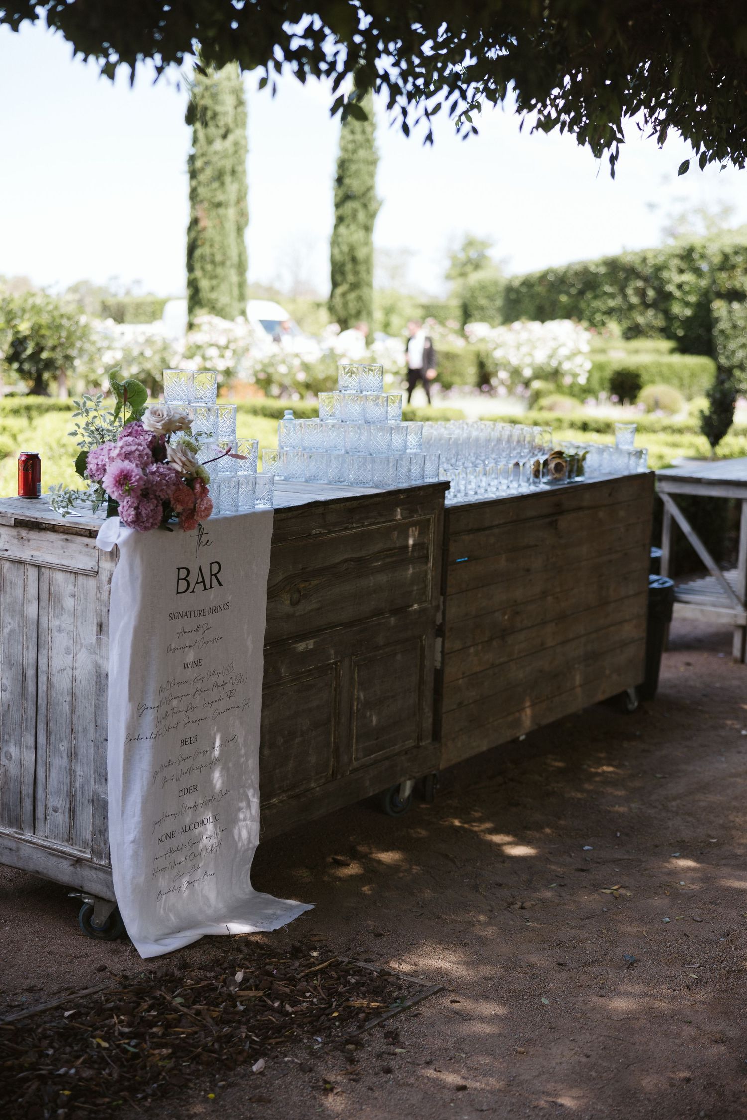 Wooden bar station decorated with pink florals and stacked glassware for outdoor service.
