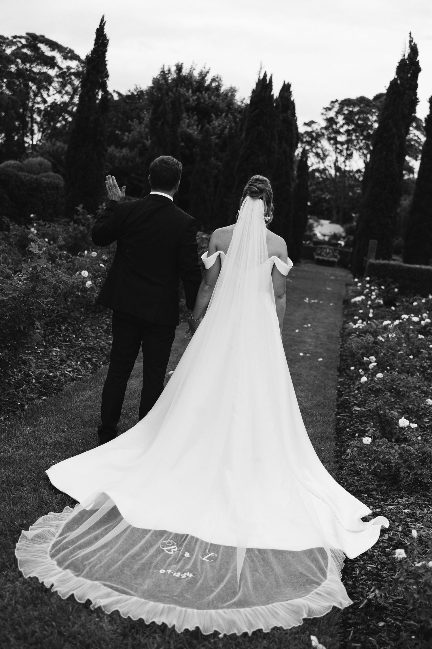 Black and white photo showcasing the dramatic trail of a wedding dress in a garden.