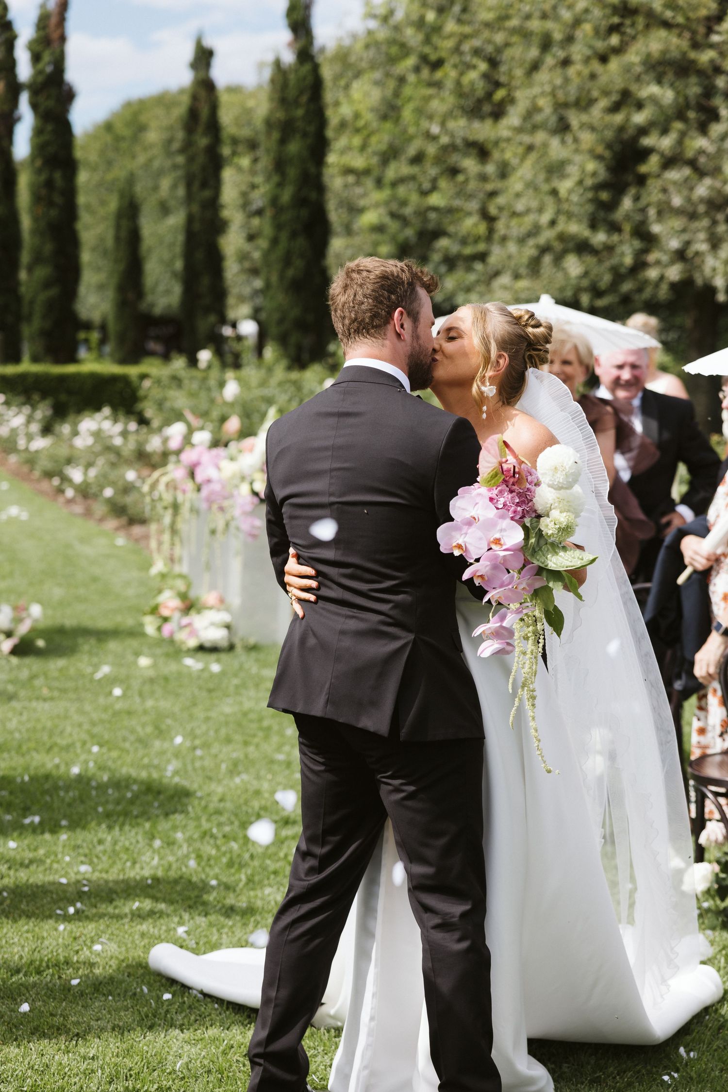 Intimate wedding moment captured in garden with tall cypress trees and pink flowers.