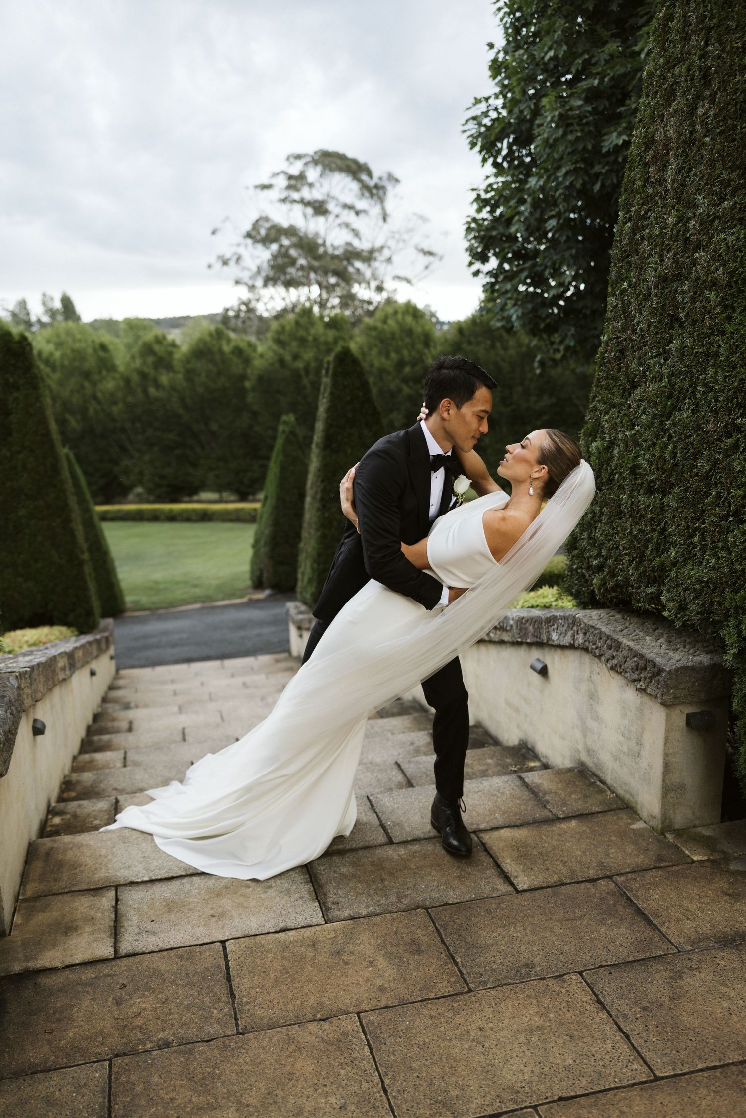 Romantic dip pose of newlyweds among sculpted garden hedges on stone terrace.