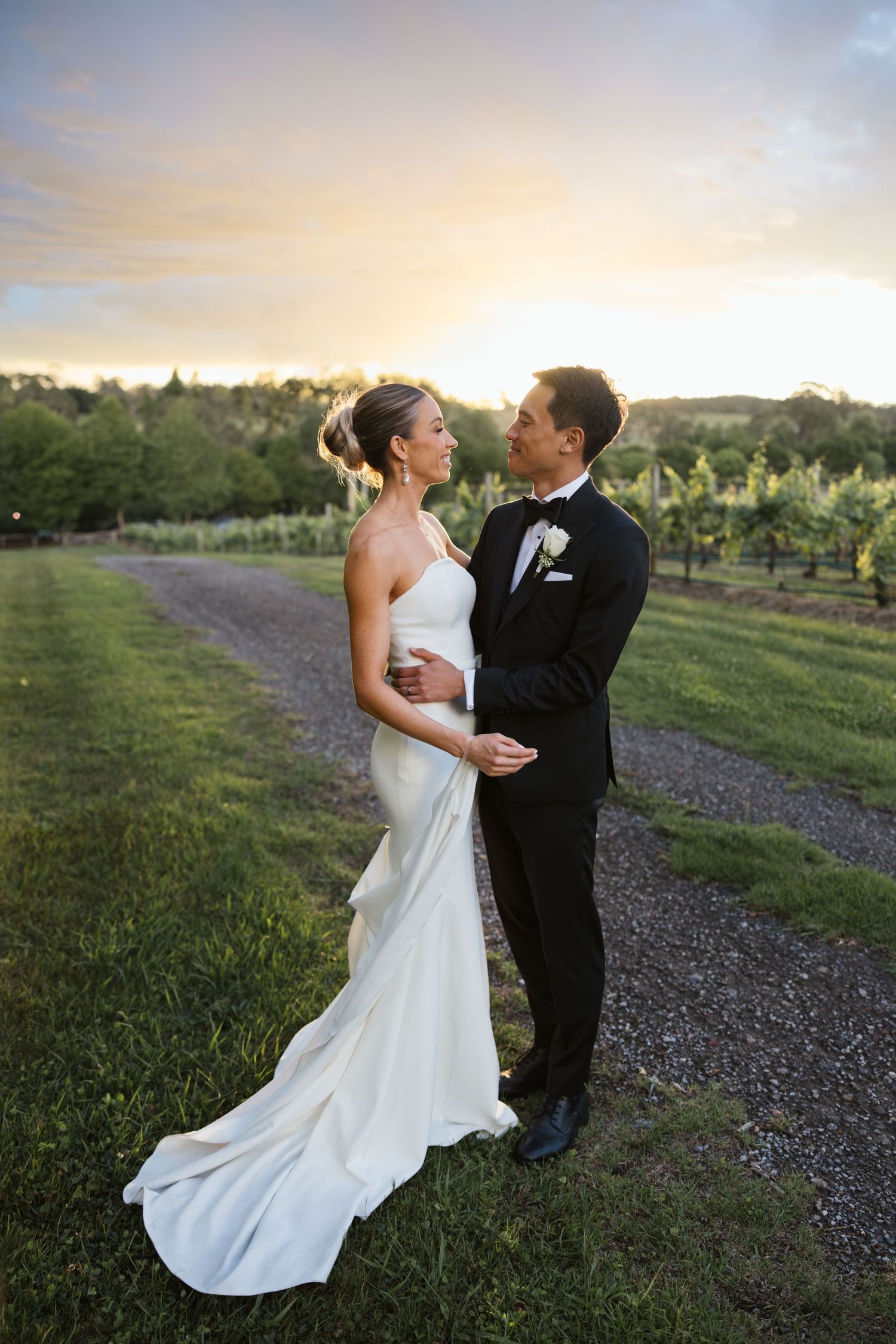 A couple embraces on a gravel path overlooking a vineyard at sunset.