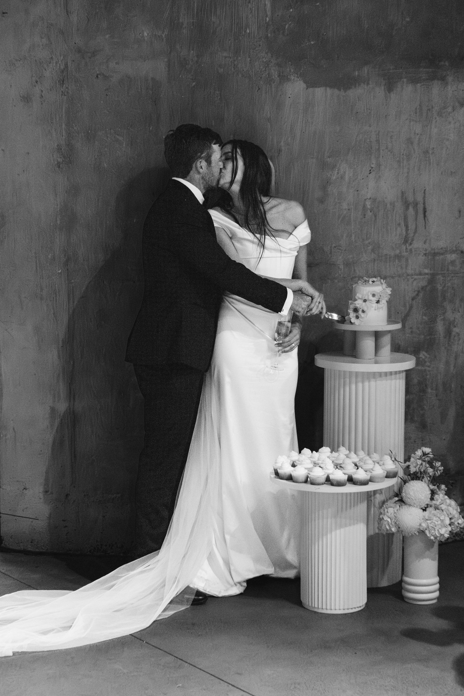 Black and white portrait of couple by wedding cake display in industrial setting.