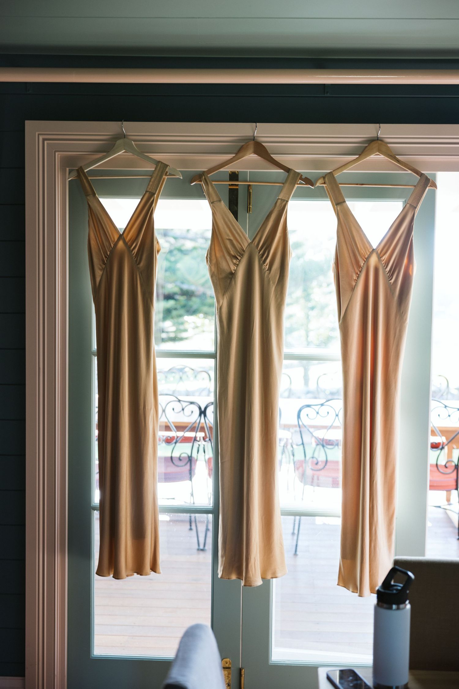 Three champagne-colored bridesmaid dresses hanging by window in elegant room.