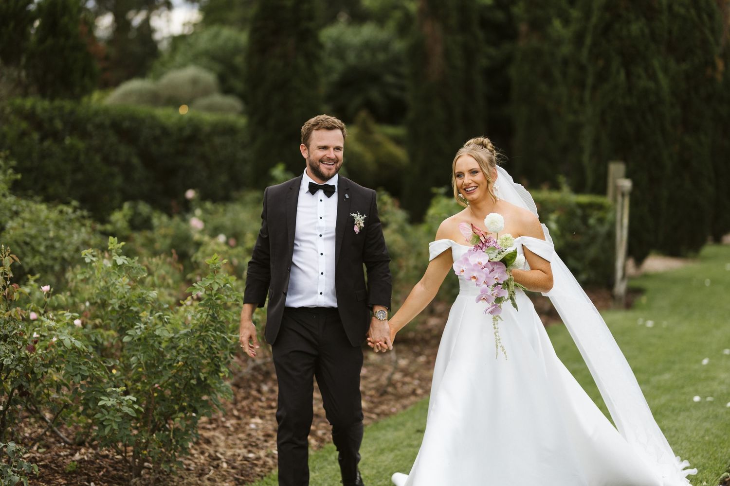 A couple poses with joy on a lush garden path during their wedding day.