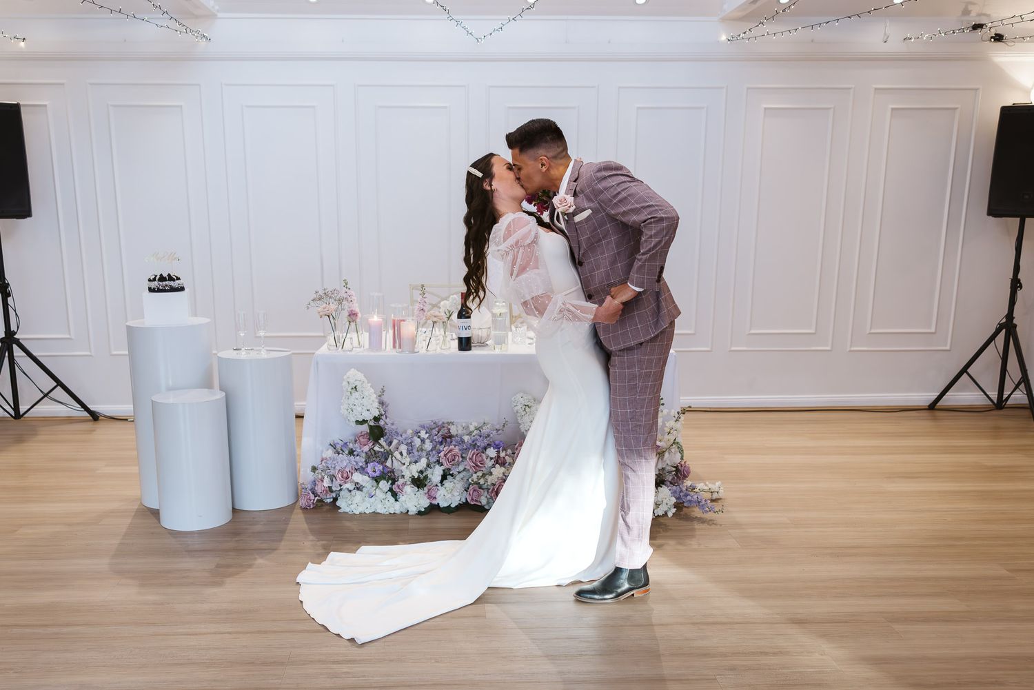 Bride and groom share a romantic first dance kiss in an elegant white ballroom with floral decorations.