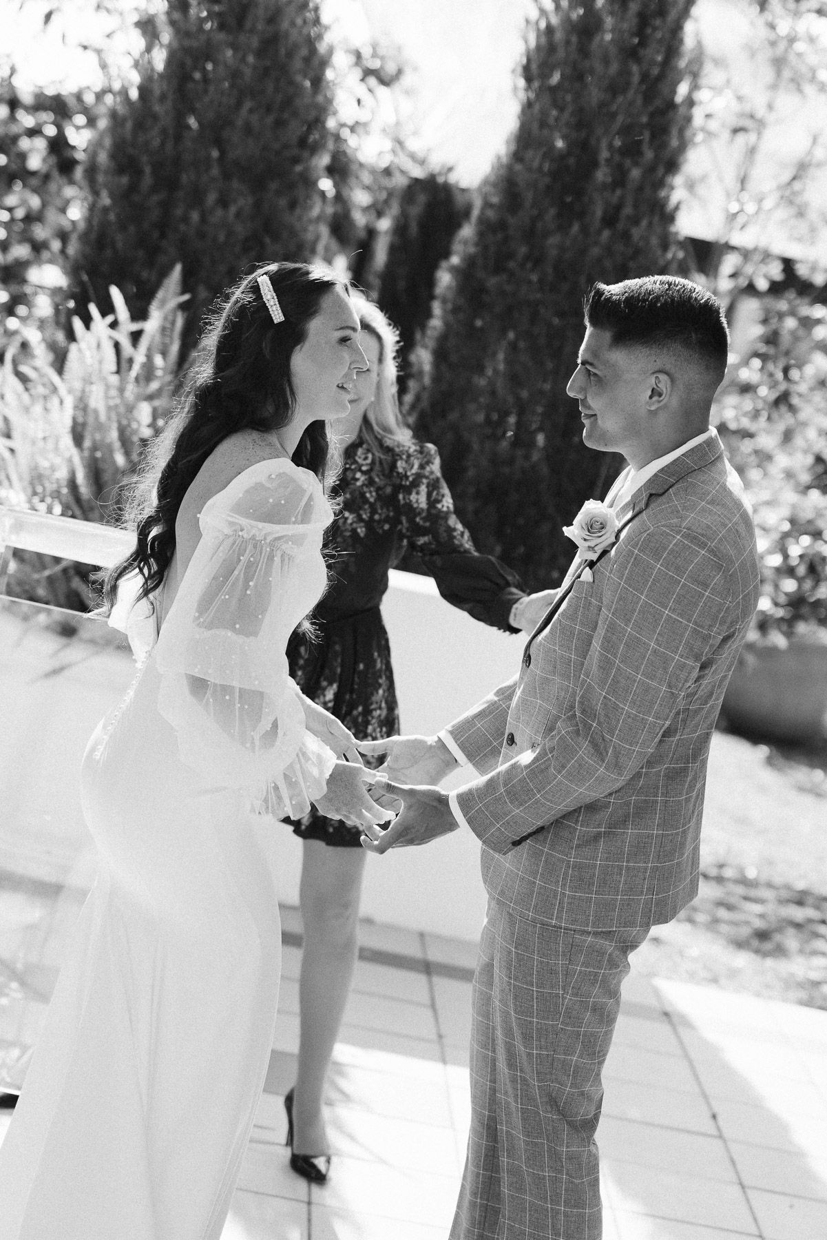 Black and white photo of a bride and groom holding hands during an outdoor wedding ceremony with an officiant.