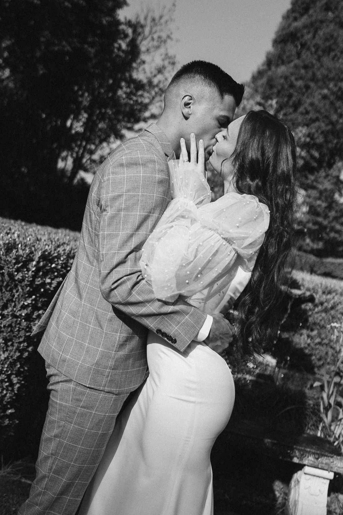 Black and white photo of a couple in elegant attire sharing a romantic moment in a lush garden setting.