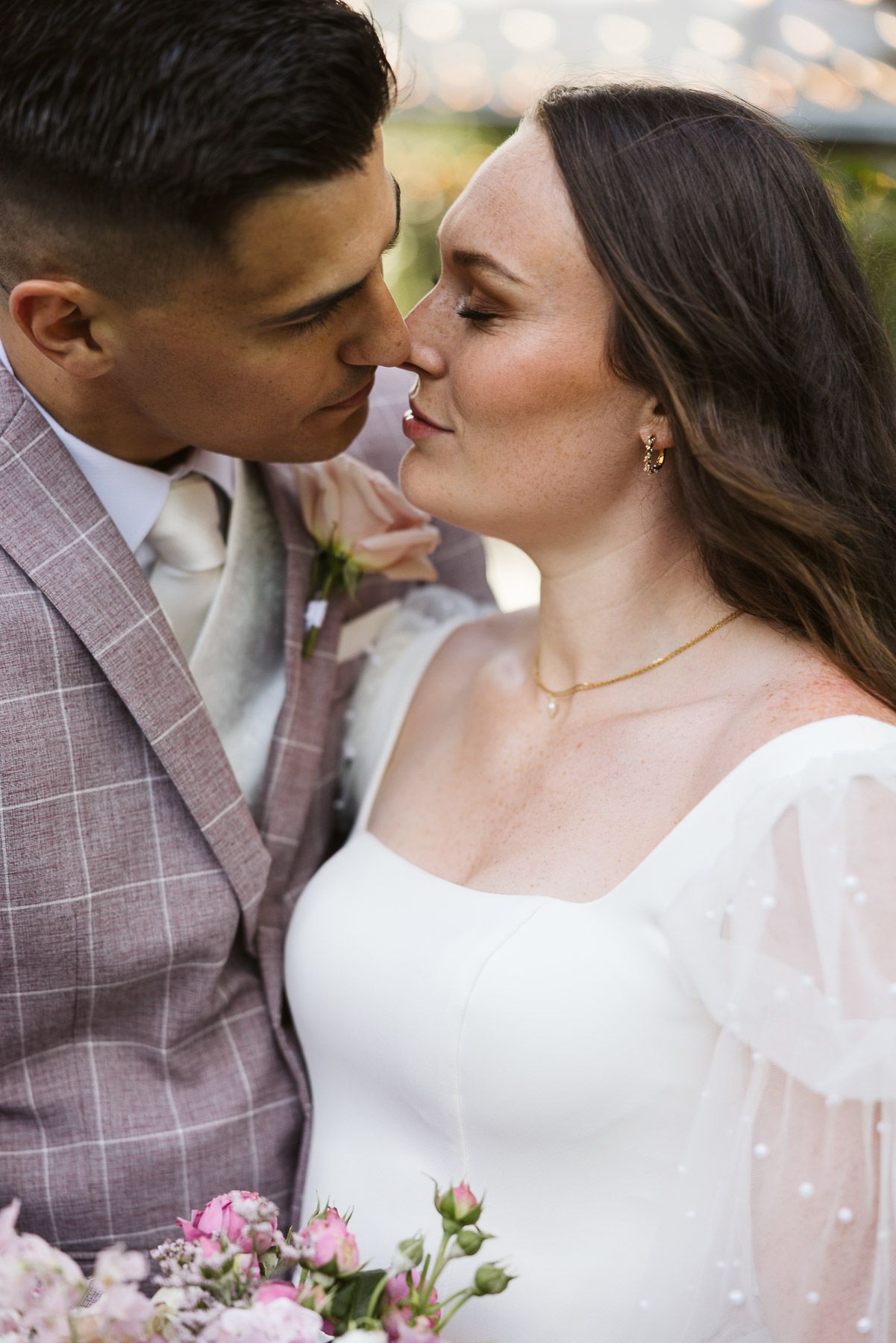 A bride and groom share a romantic almost-kiss, wearing wedding attire with pink floral bouquet.