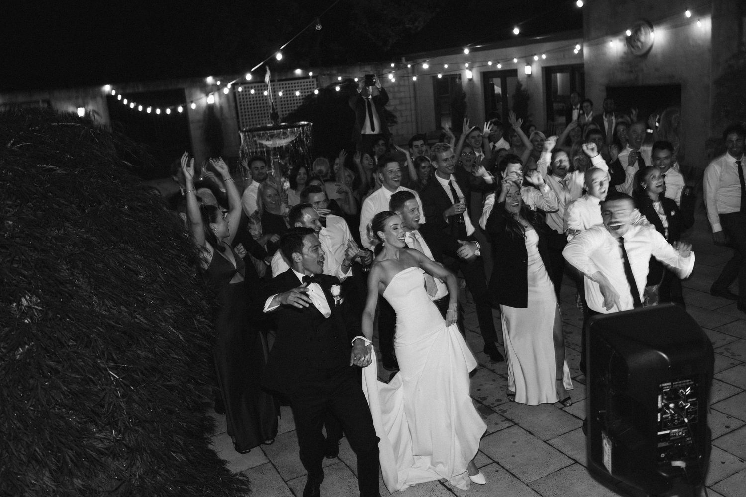 A black and white photo of wedding guests dancing under string lights at an outdoor reception.