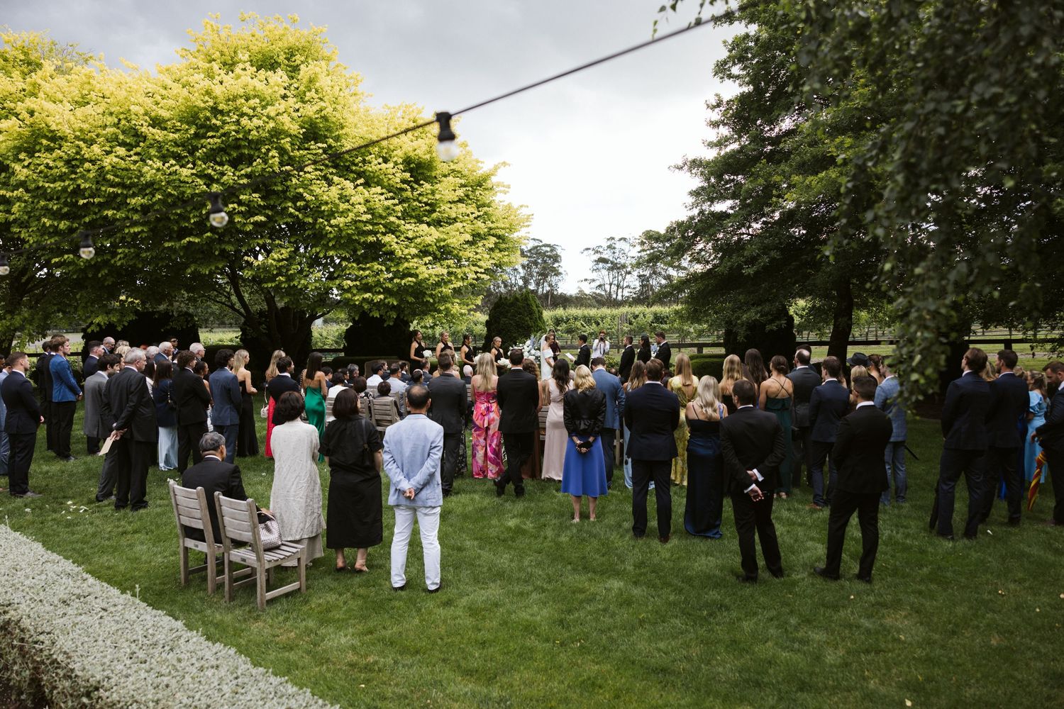 Wedding guests gathered on a lawn during an outdoor ceremony with trees in the background.
