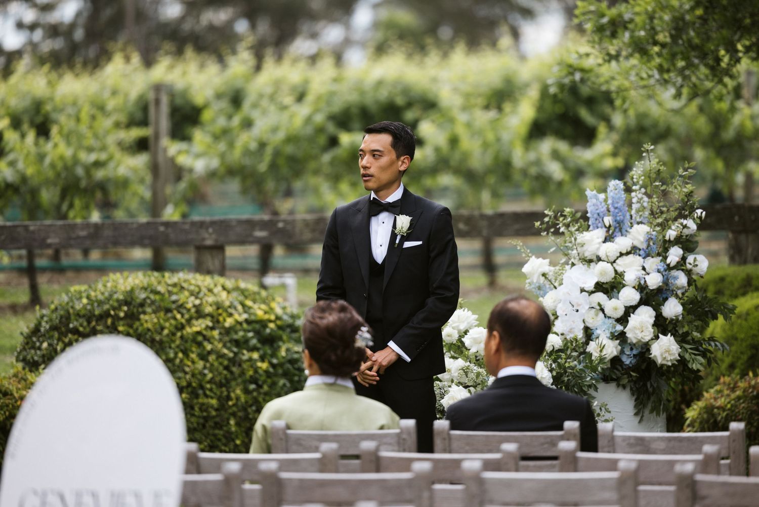 Outdoor wedding ceremony setup with white floral arrangements and guest seating in a garden setting.