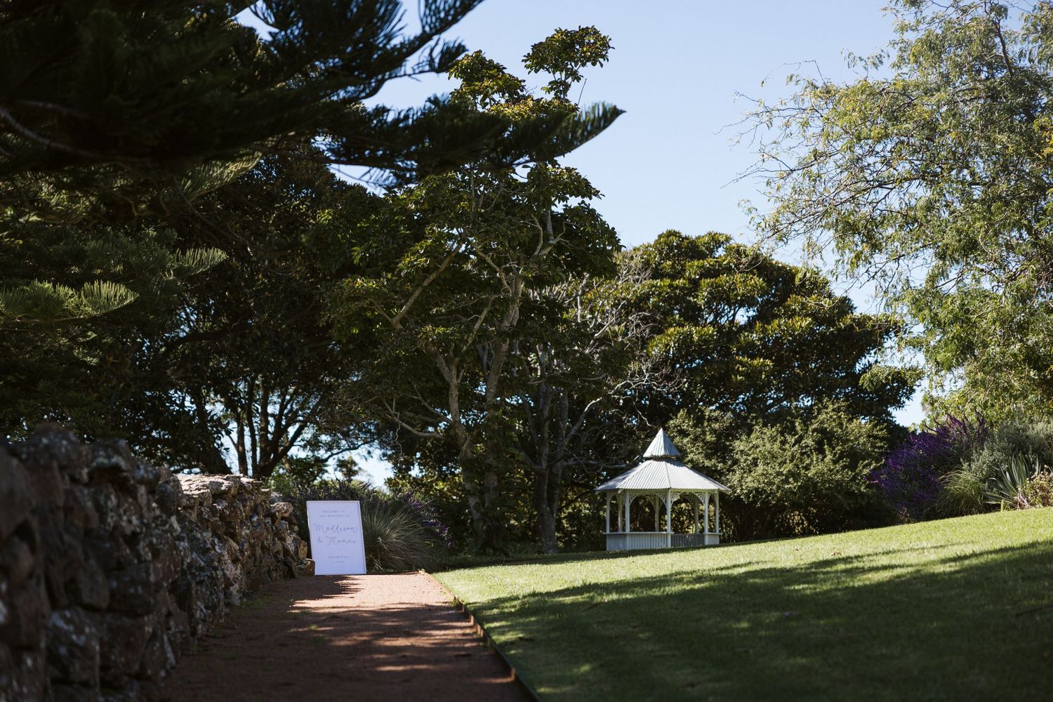 White gazebo sits on a grassy hill surrounded by trees in a serene garden setting.