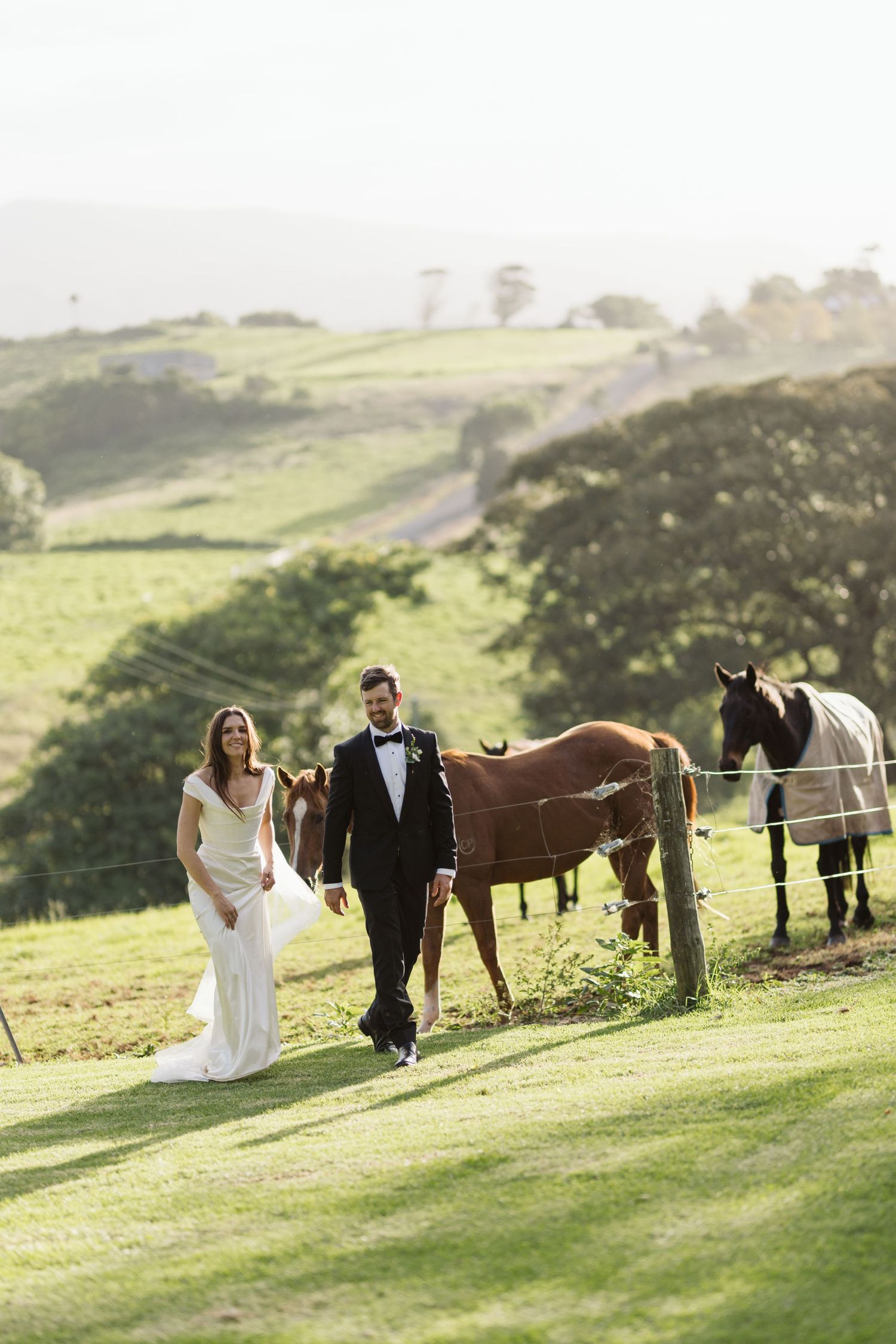 Newlyweds walking together on grassy hillside with horses grazing in the background.