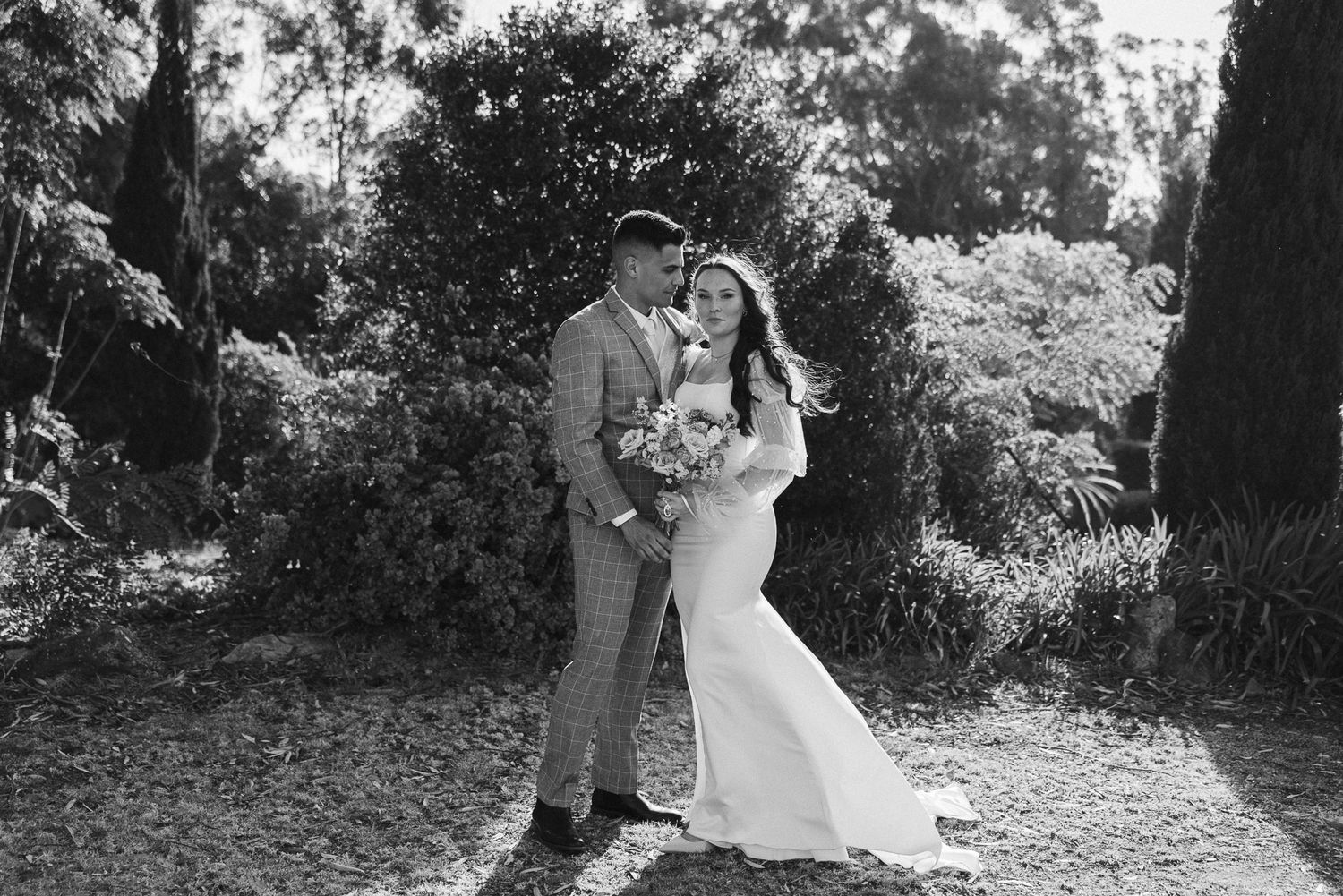 Black and white photo of a bride and groom embracing in a lush garden setting on their wedding day.