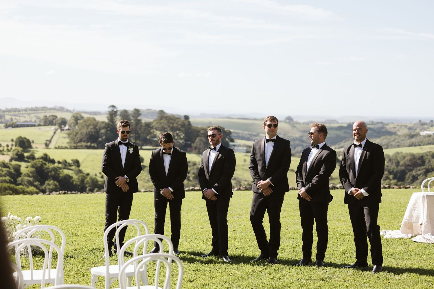 Groomsmen in black suits standing together on hilltop overlooking countryside.