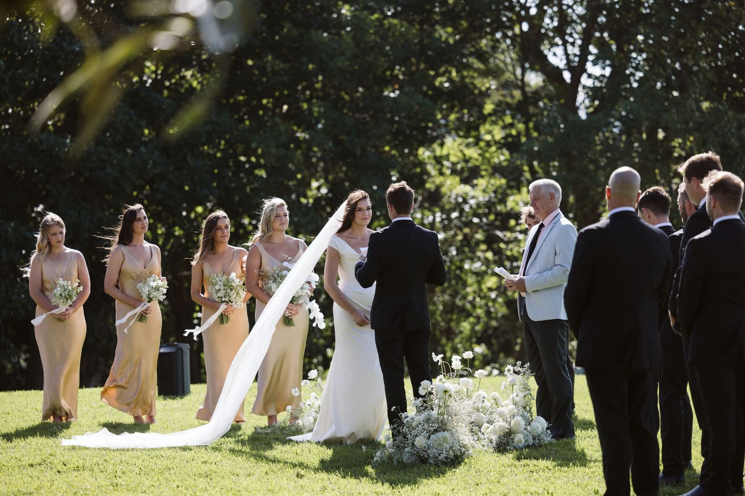 Wedding ceremony taking place outdoors on a sunny day with bridal party and guests gathered on a lush green lawn.