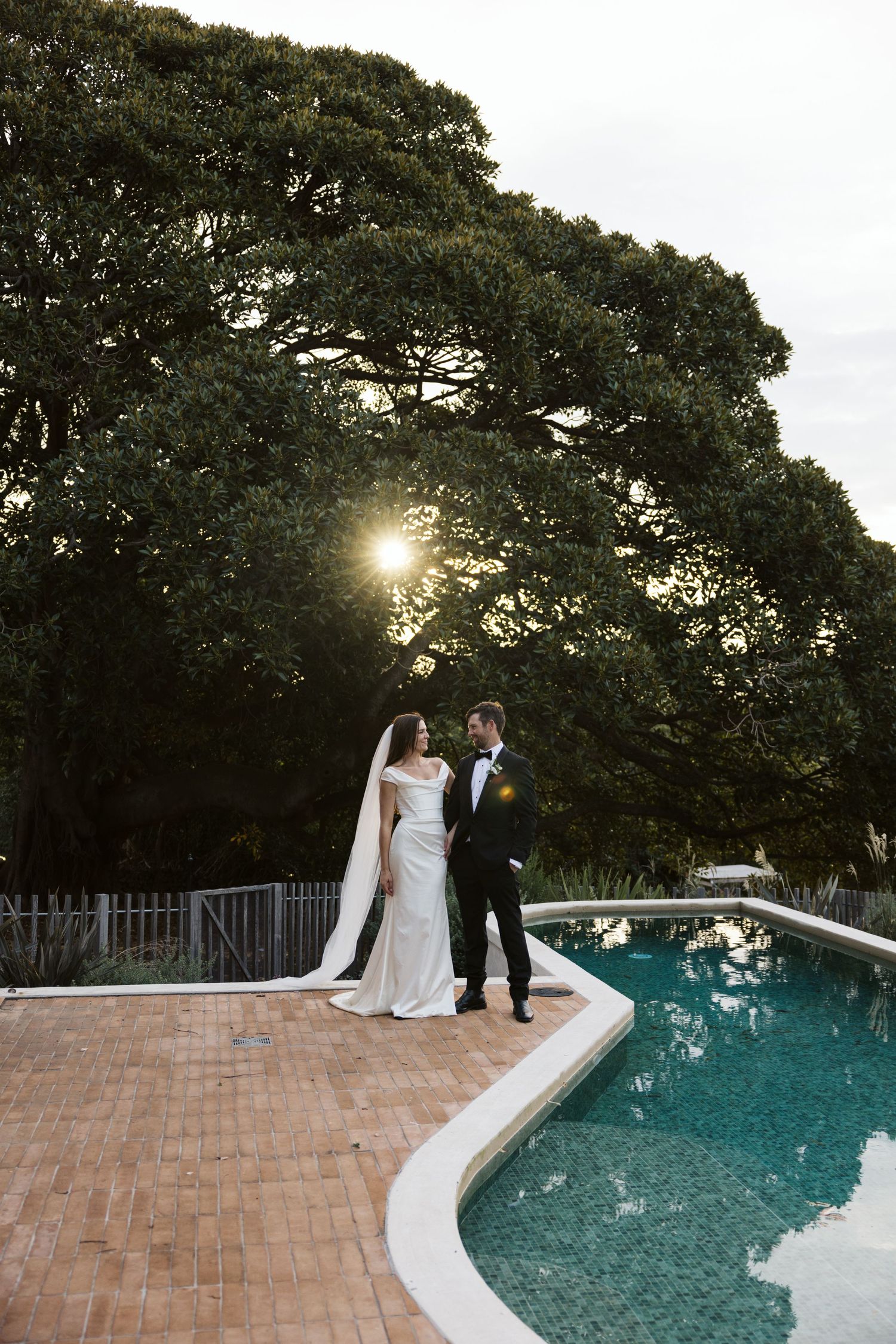 Wedding couple stands by a poolside at sunset with large trees creating a dramatic silhouette background.