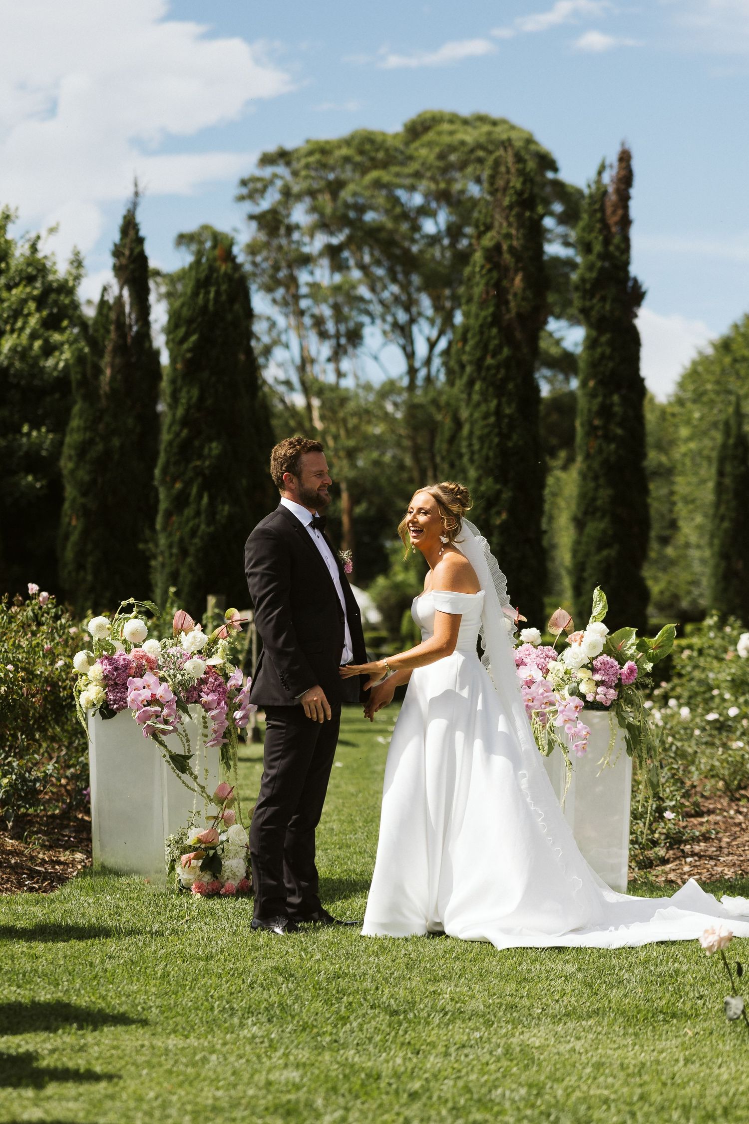 Romantic wedding ceremony in a manicured garden with tall cypress trees and floral arrangements.