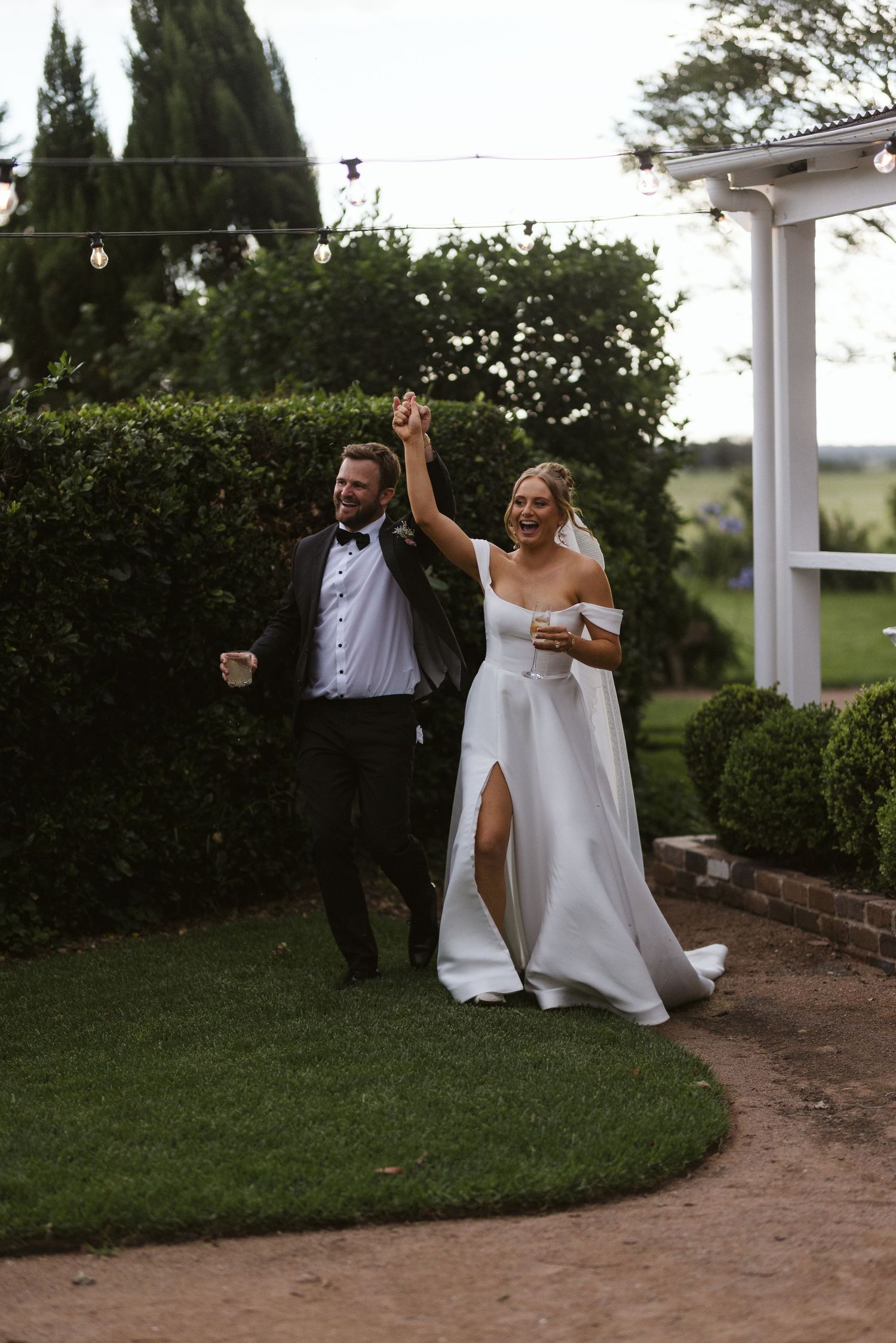 Joyful couple celebrates with raised arms on garden path during wedding.