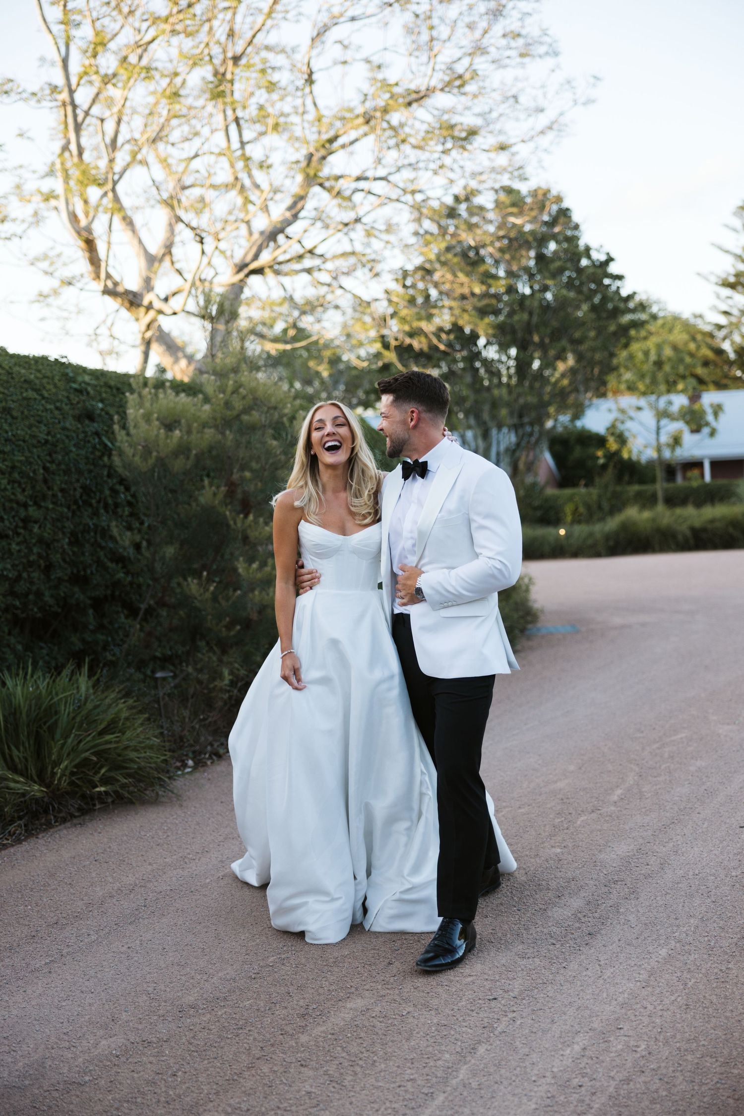 A joyful wedding couple walks along a garden path, with the bride wearing an elegant white dress.