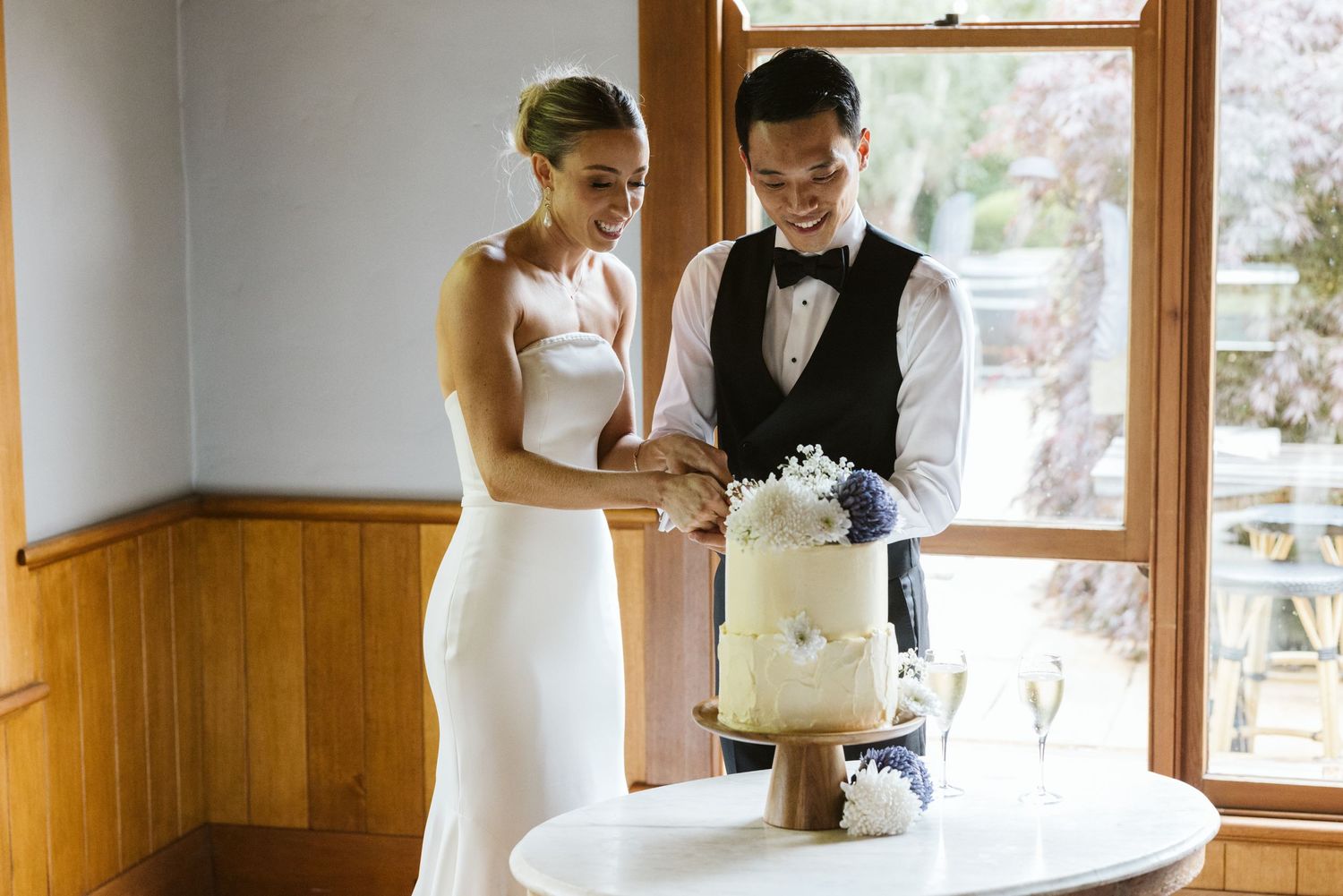A couple cuts their elegant white wedding cake together by a window.