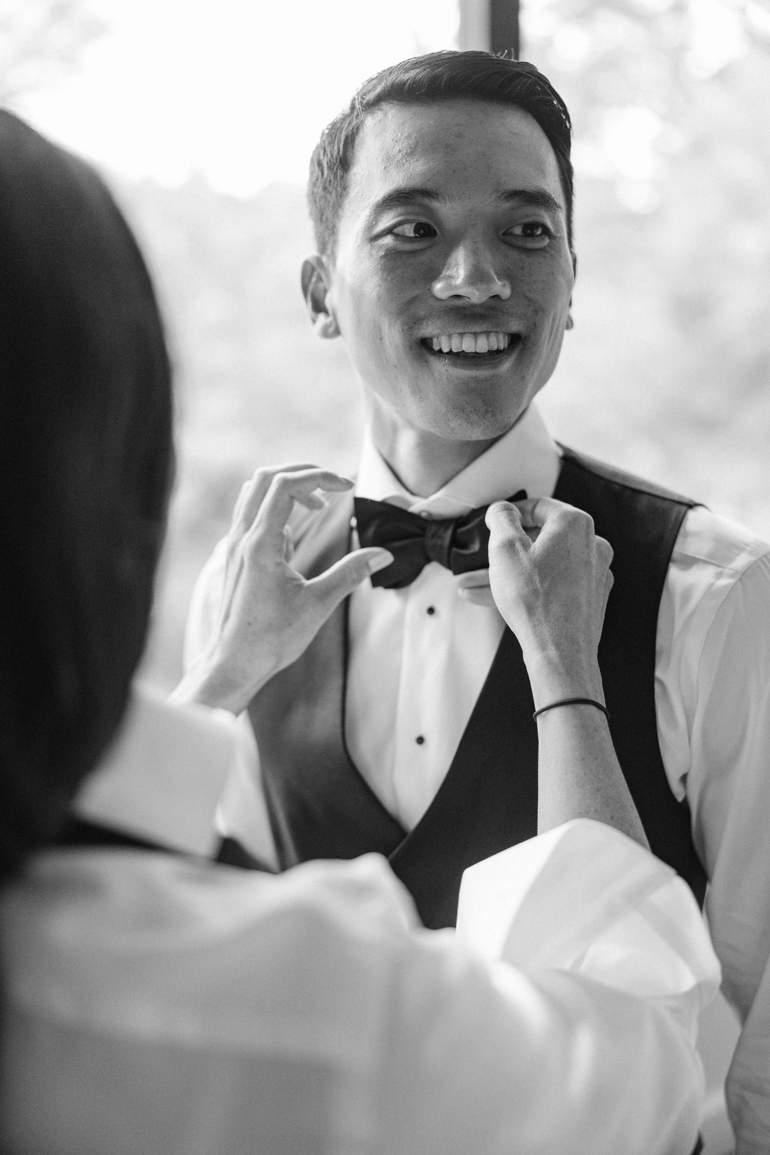 A joyful groom in a tuxedo vest smiles while someone adjusts his bowtie on his wedding day.