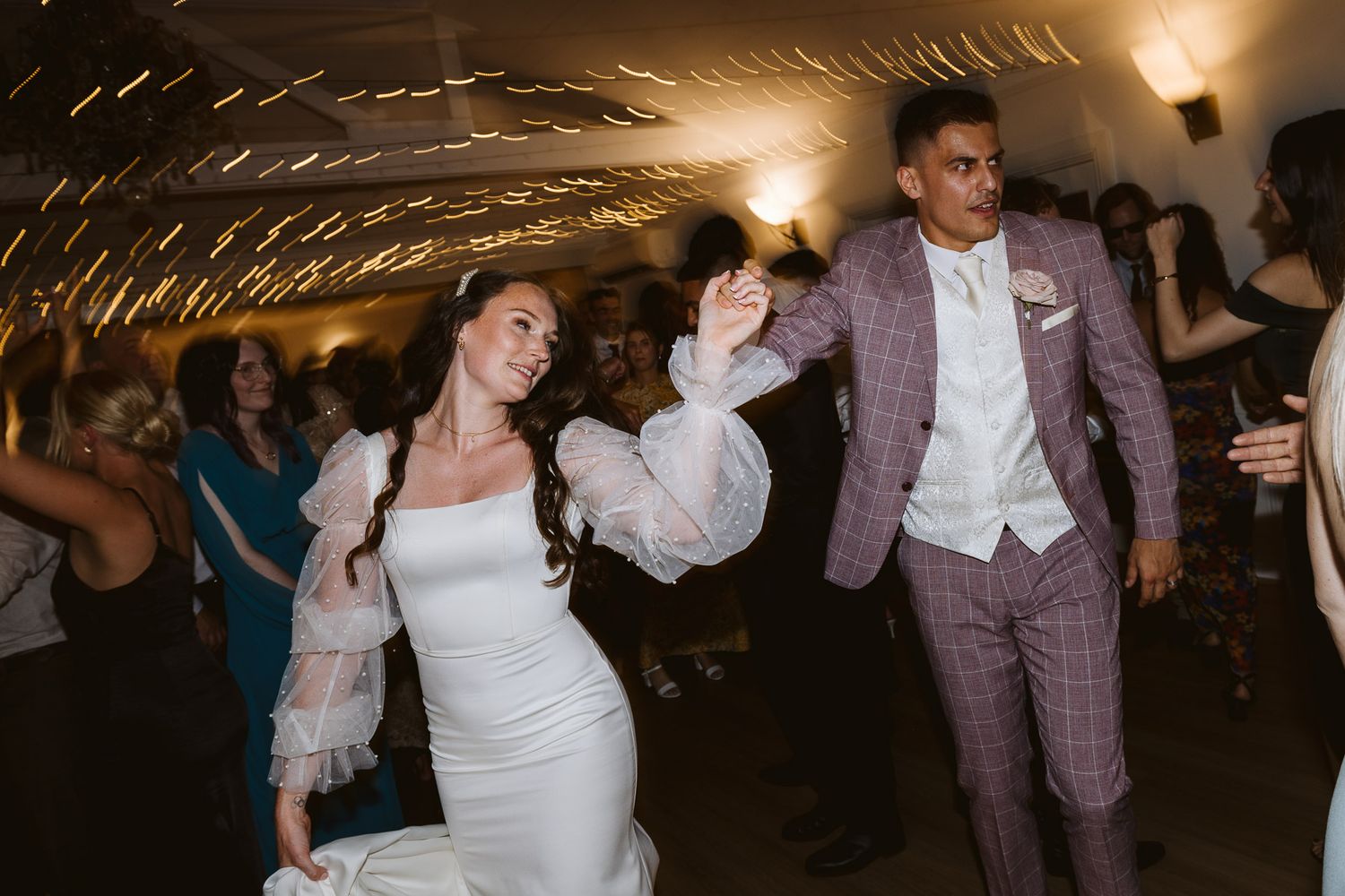 Bride in white gown and groom in plaid suit dancing joyfully at wedding reception with sparklers.