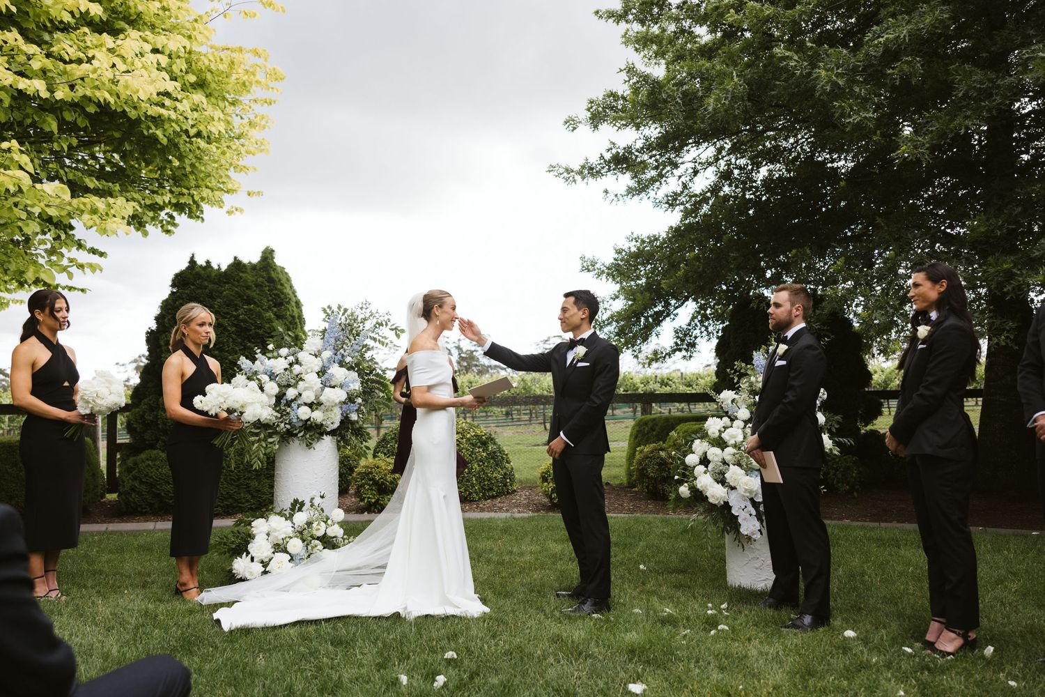 Wedding ceremony taking place outdoors with attendants in black attire and white floral decorations.