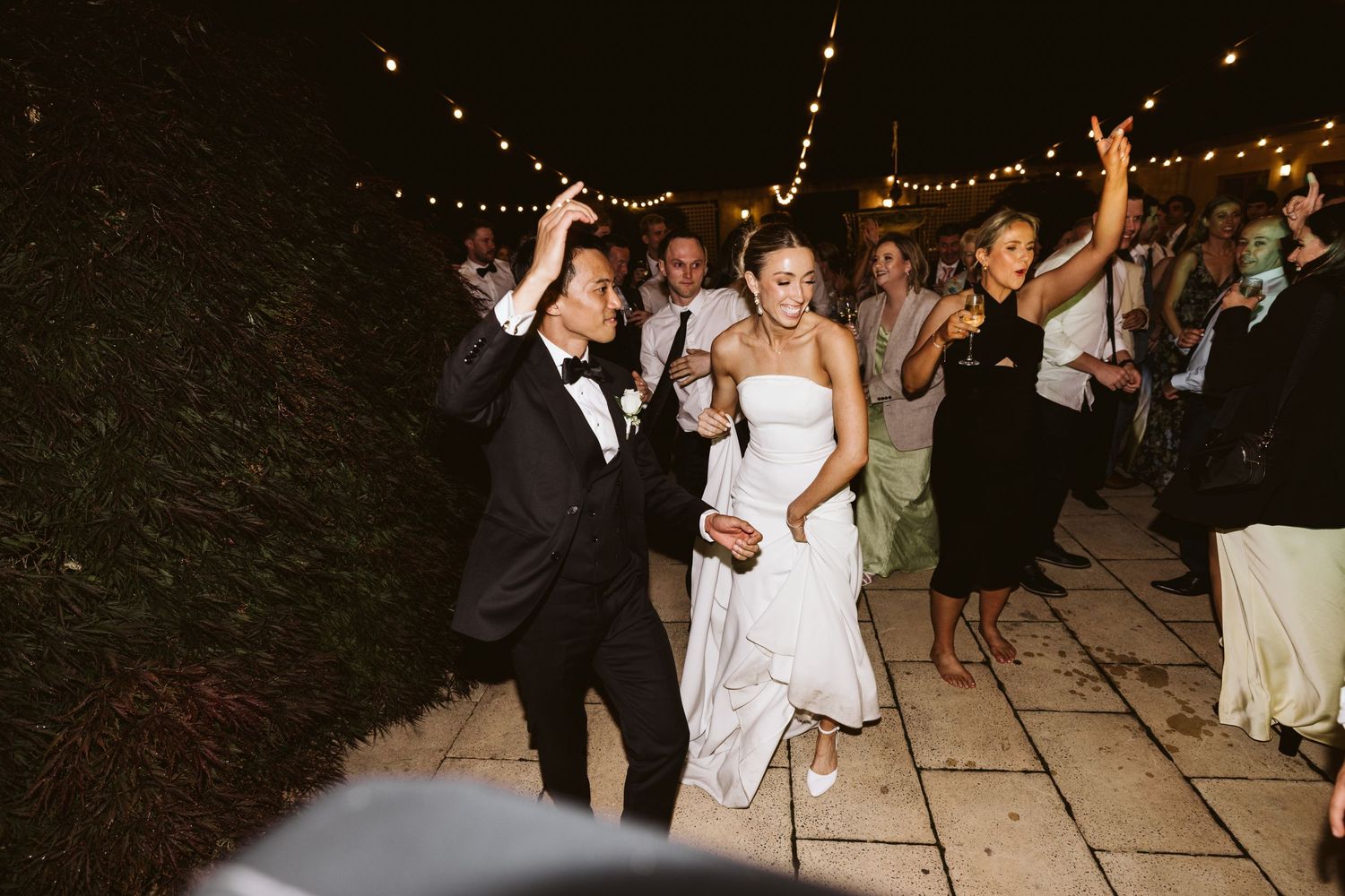 Wedding guests dancing and celebrating on an outdoor patio with string lights at night.