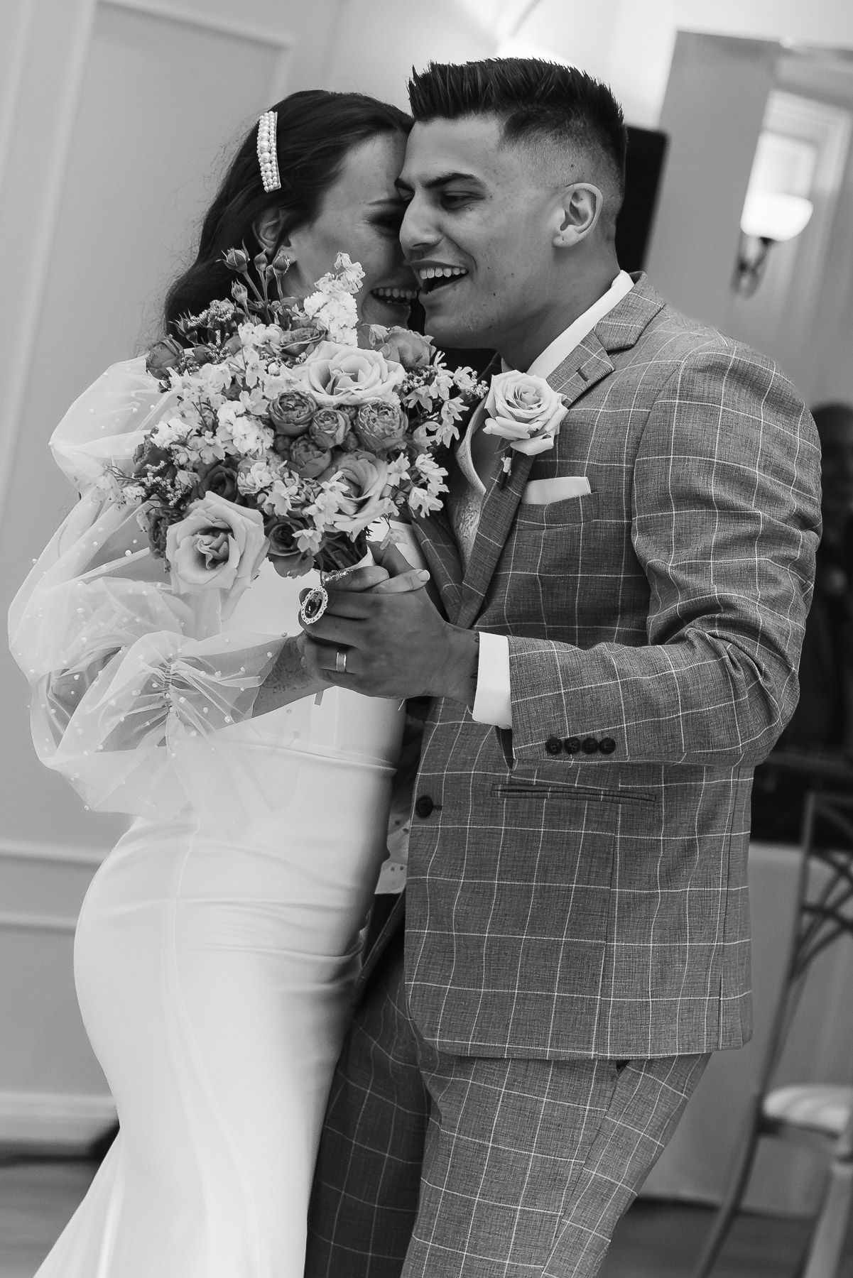 Black and white photo of a laughing bride and groom embracing, holding a floral bouquet on their wedding day.