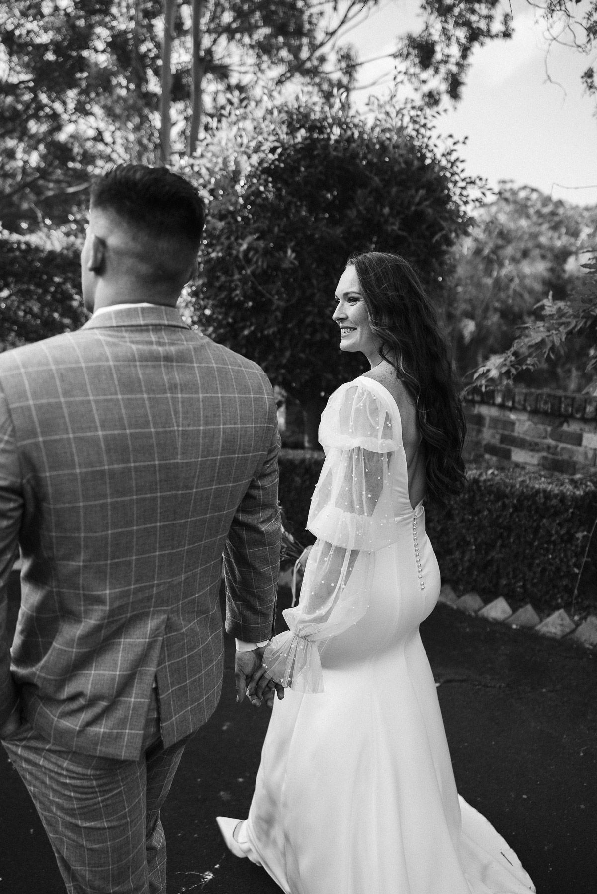 Black and white photo of a bride and groom holding hands, walking outdoors, bride in white gown glancing back smiling.