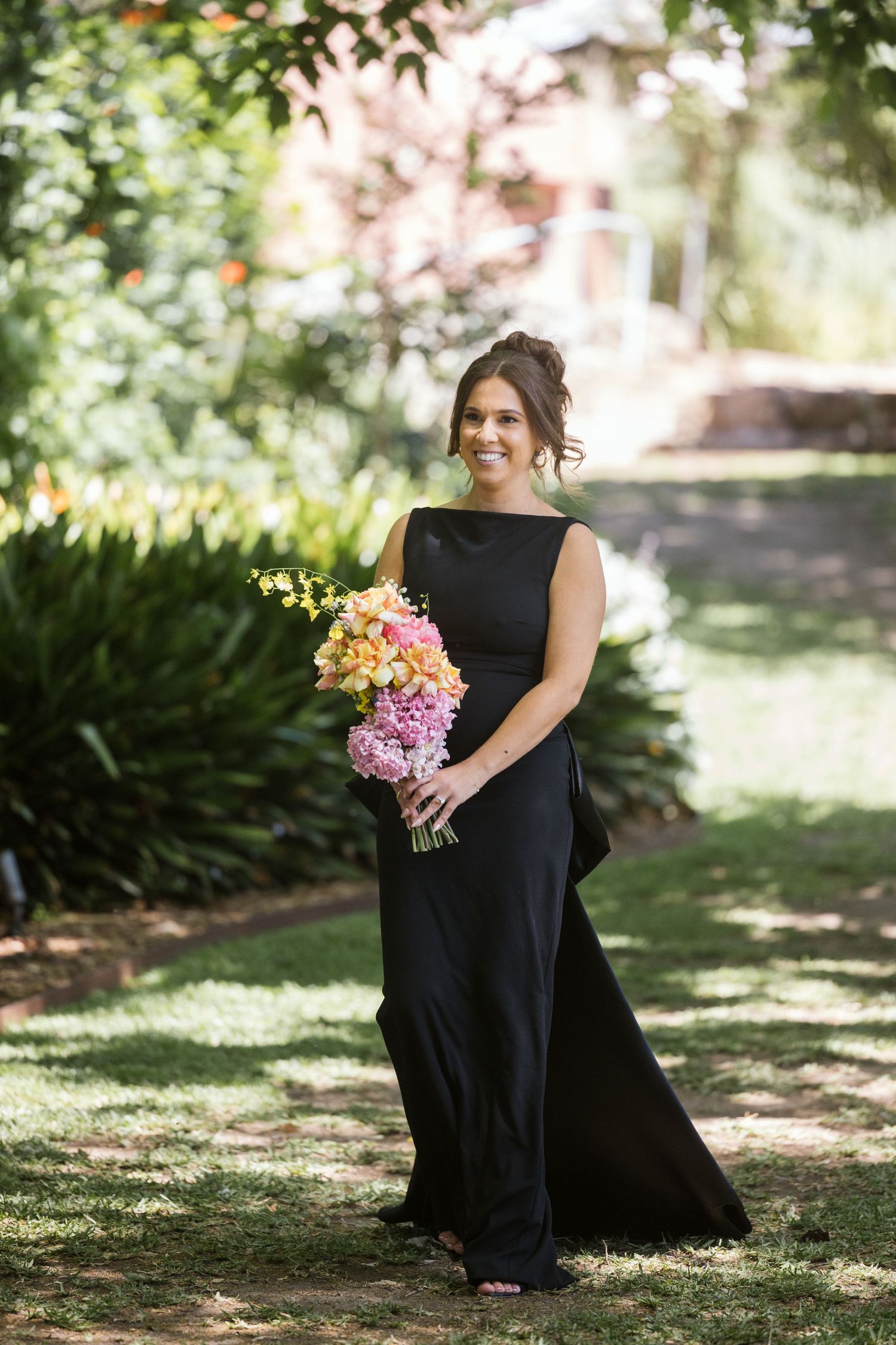 Bridesmaid poses with pink and yellow bouquet in garden wearing classic black gown.