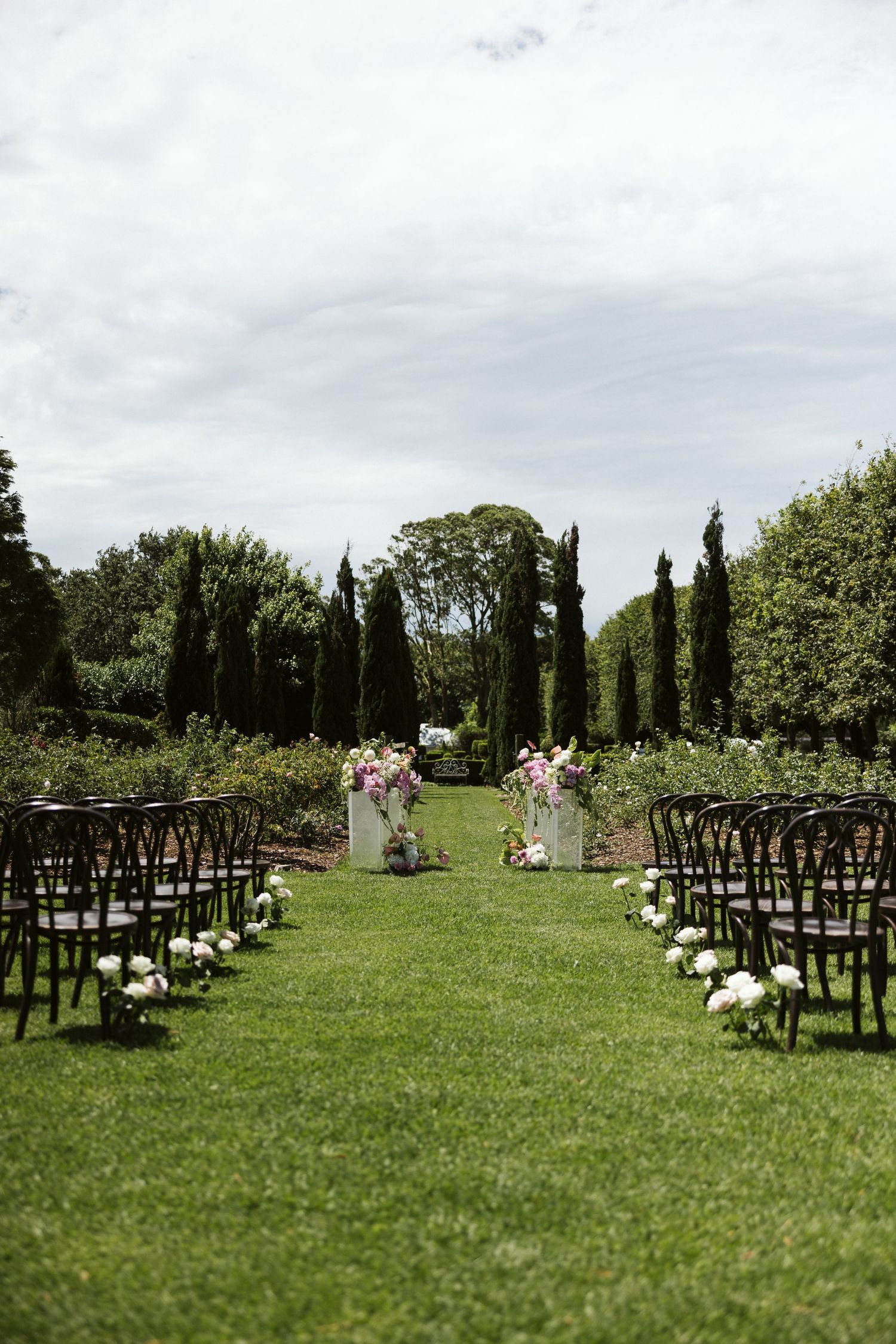 An outdoor wedding ceremony setup with rows of dark chairs and floral arrangements on a manicured lawn.