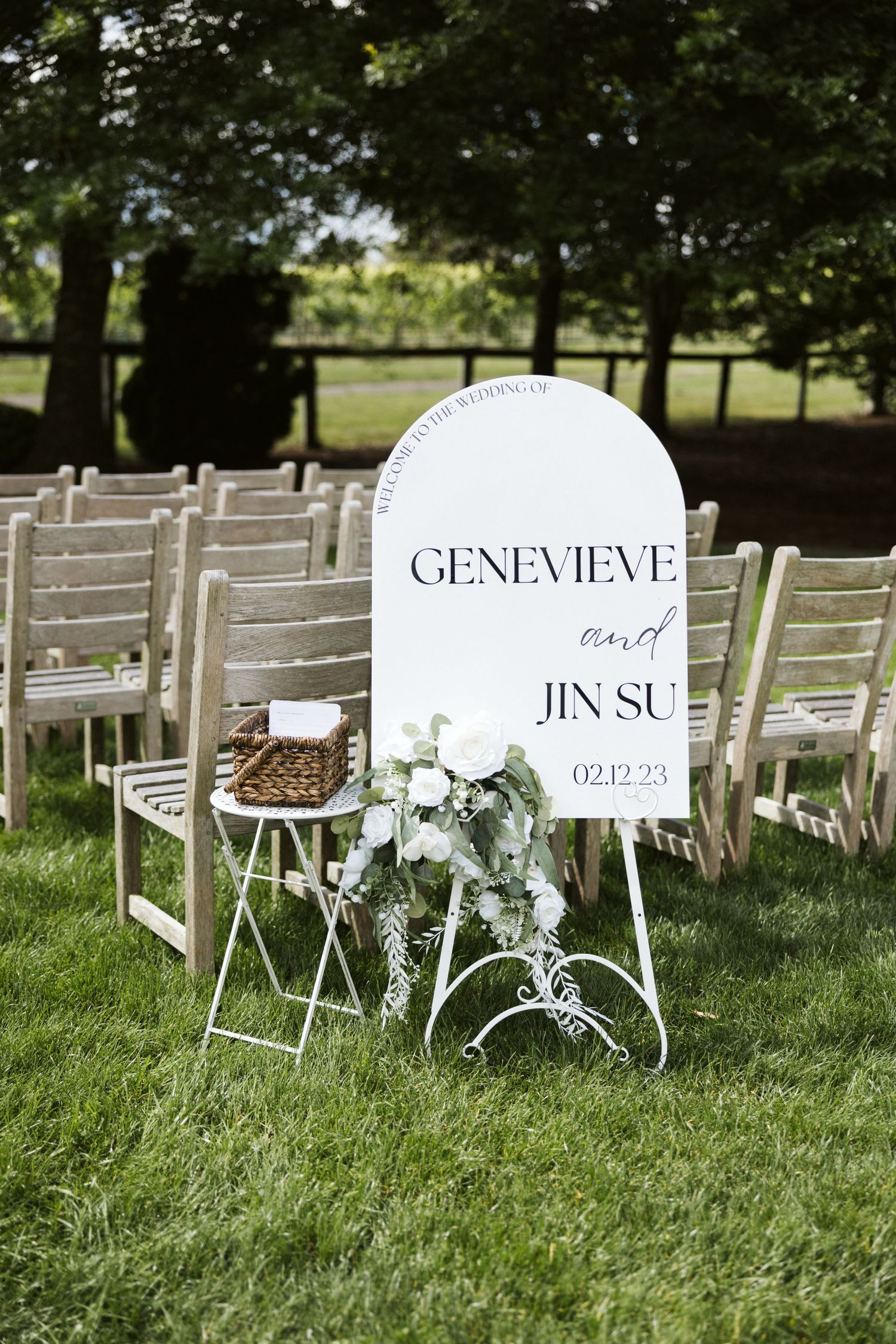 A wedding welcome sign with floral decorations displayed in front of white chairs outdoors.