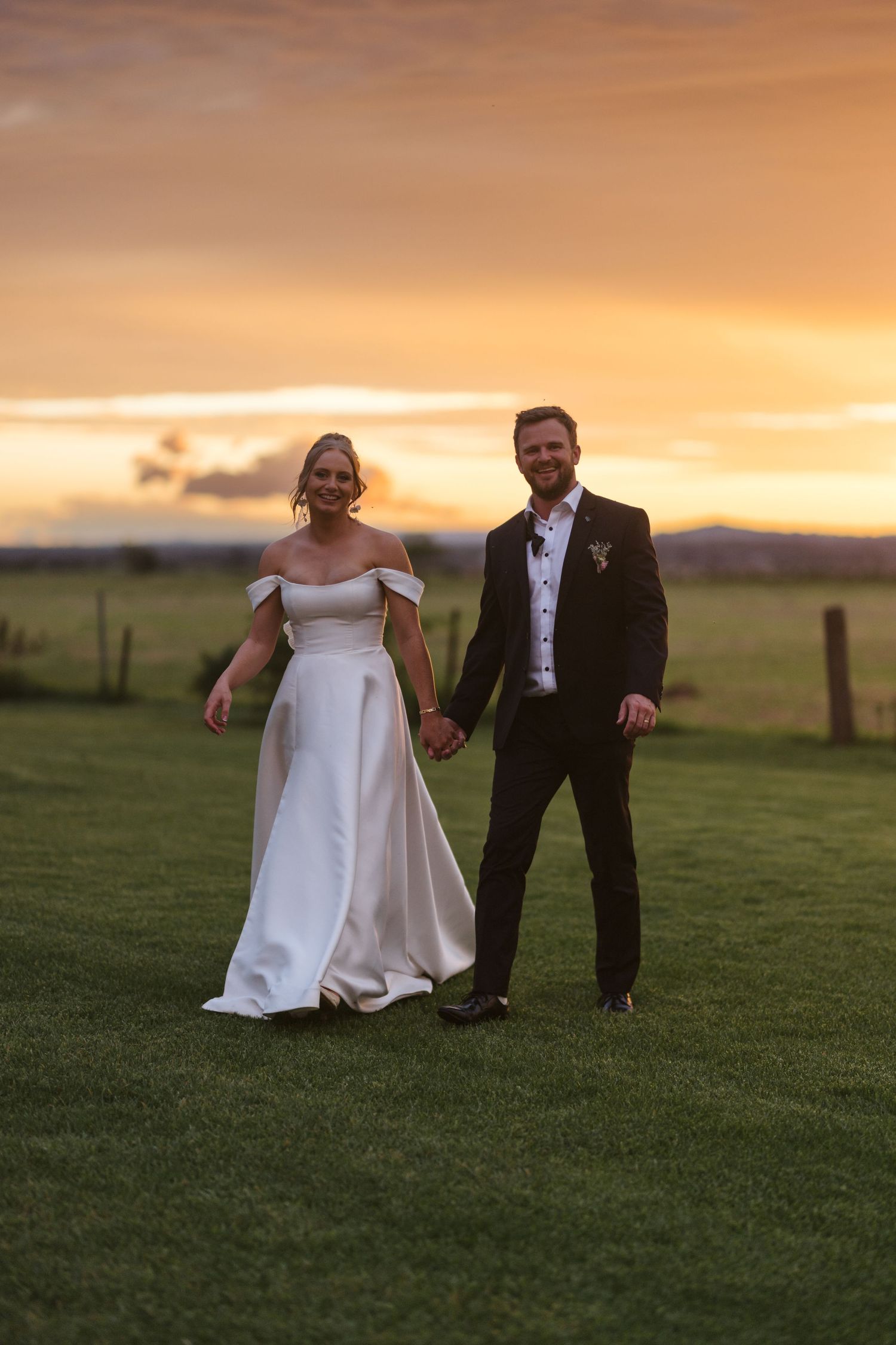 Sunset wedding portrait with couple standing together on grassy field with mountains in background.