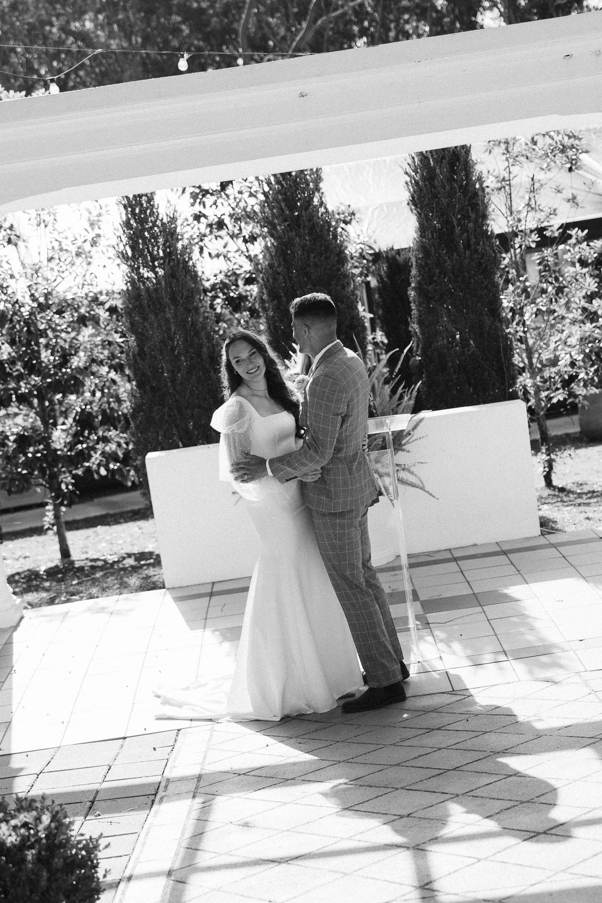 Black and white photo of a bride and groom sharing a tender embrace outdoors under a white pergola.