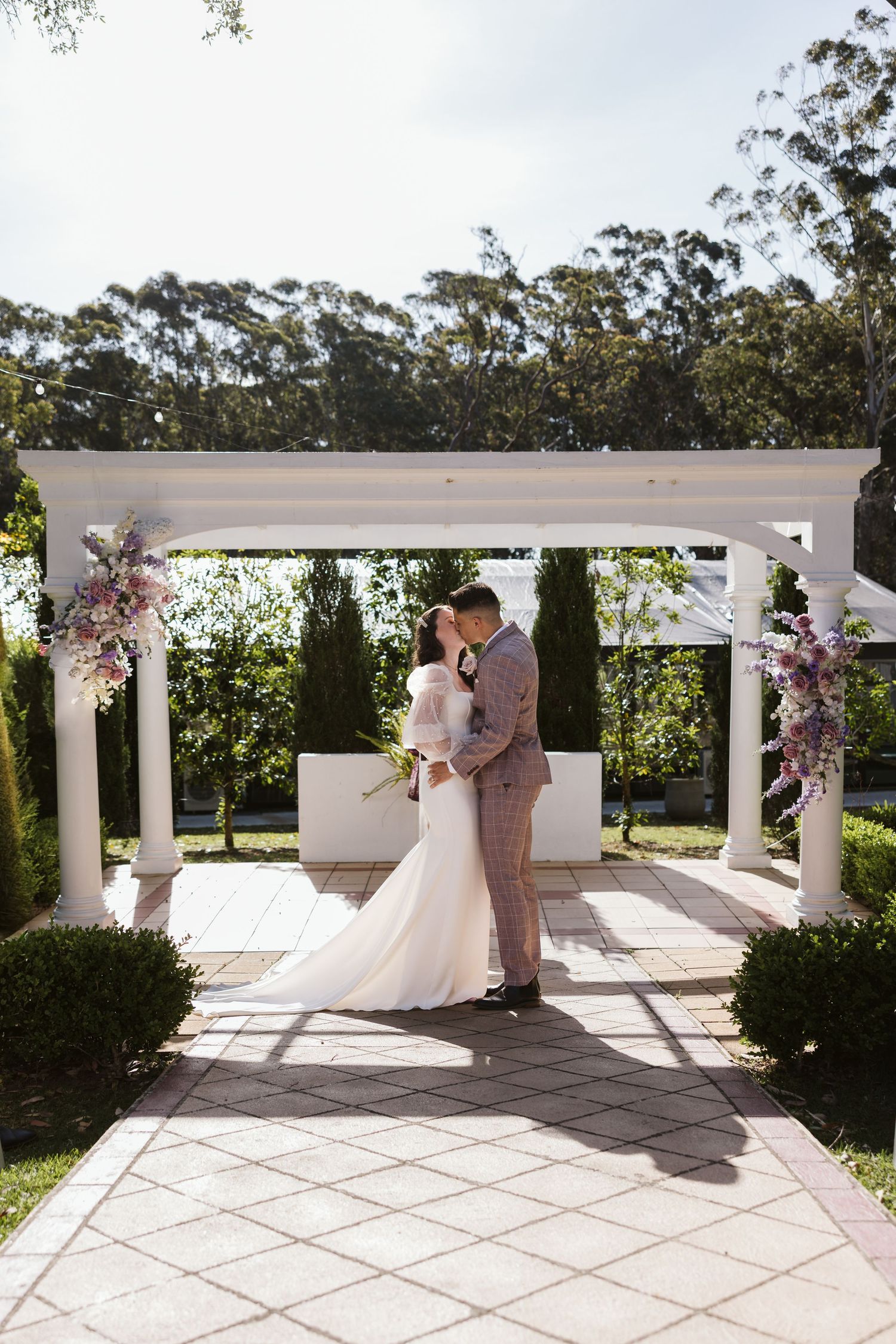 A couple shares a romantic moment under an elegant white pergola decorated with floral arrangements in a garden setting.