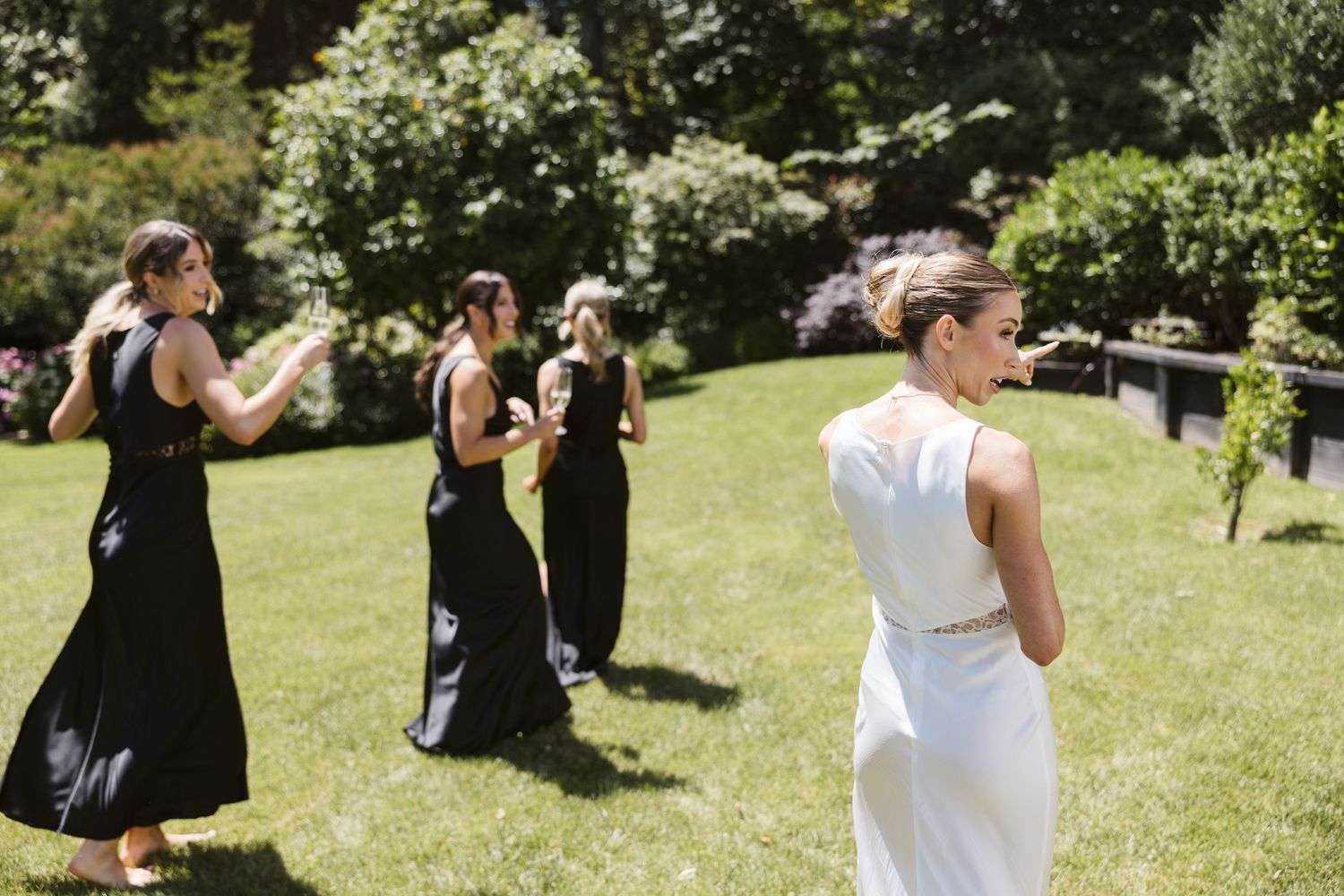 Bridesmaids in black dresses following bride in white through sunny garden setting.