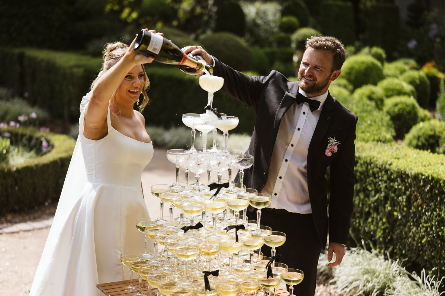 Couple pours champagne into tower of glasses at outdoor wedding celebration.
